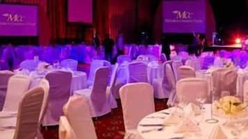 A banquet hall with round tables set for a formal event, featuring a stage with a large screen displaying the event's name.