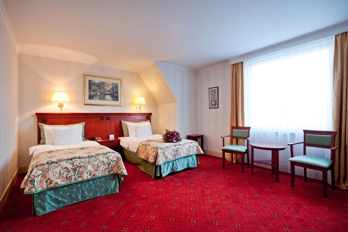 A hotel room with two beds, a sitting area with chairs, a large window with curtains, and a painting on the wall.