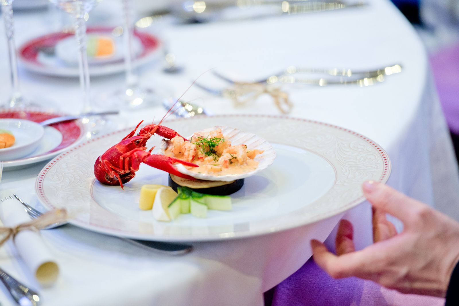 A lobster dish on a plate with lemon wedges and cucumber slices, set on a table with a white tablecloth and silverware.