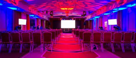 A conference room with rows of chairs, a stage with a large screen, and a red carpet.
