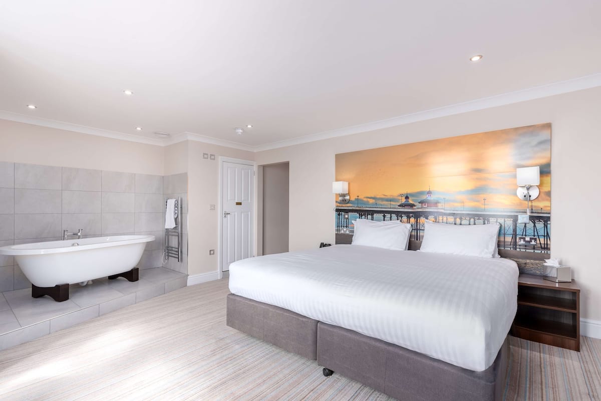A modern hotel room with a large bed, a bathtub, and a painting of a sunset over a pier.