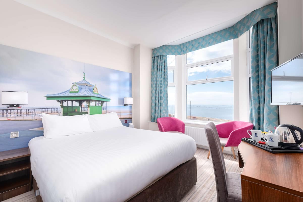 A hotel room with a large bed, a desk with a kettle, a chair, and a view of the sea.