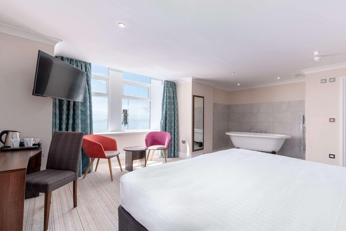 A hotel room with a bed, a TV, a desk with a coffee maker, two chairs, a window with curtains, and a bathroom with a bathtub.