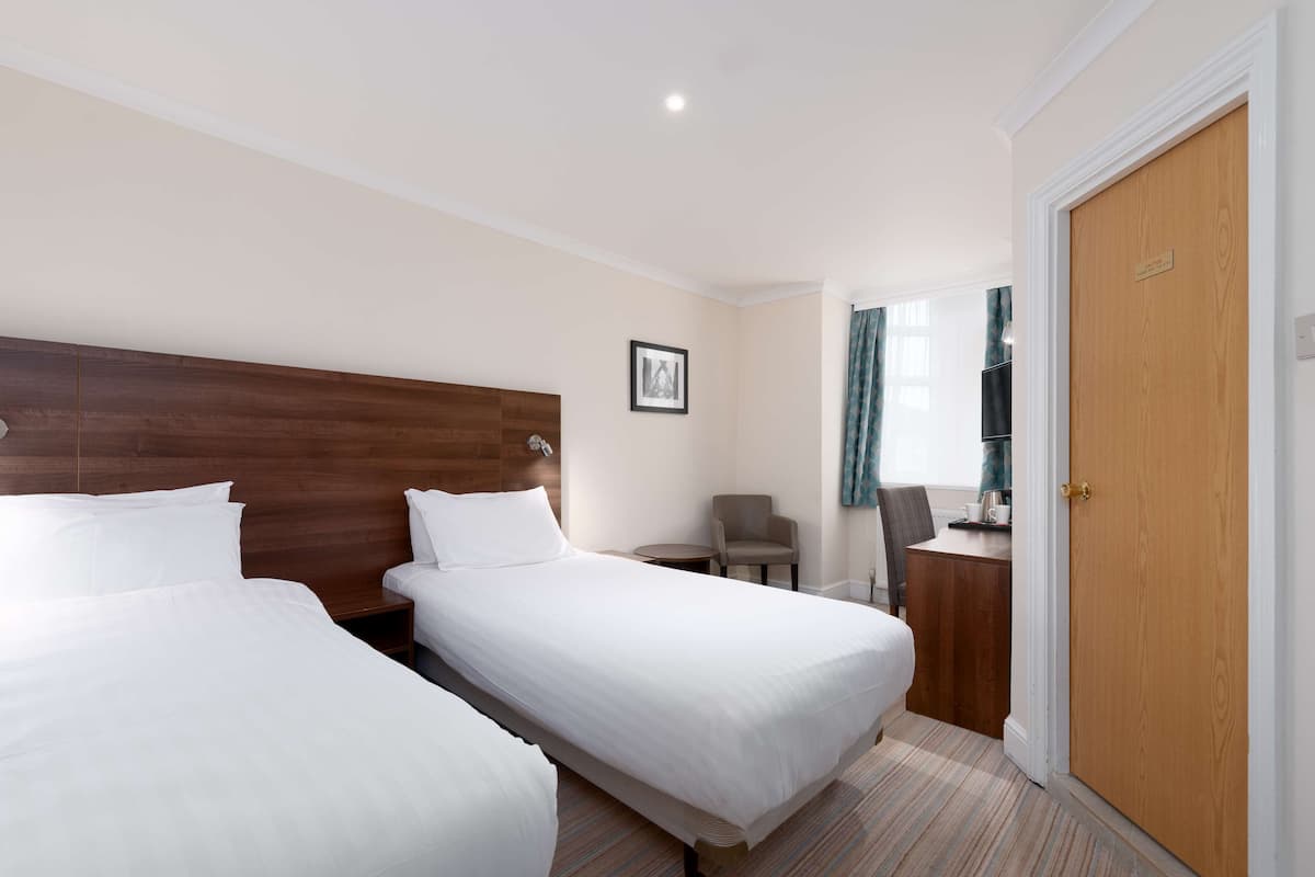 A hotel room with two single beds, a wooden headboard, a small table with a chair, and a wardrobe.