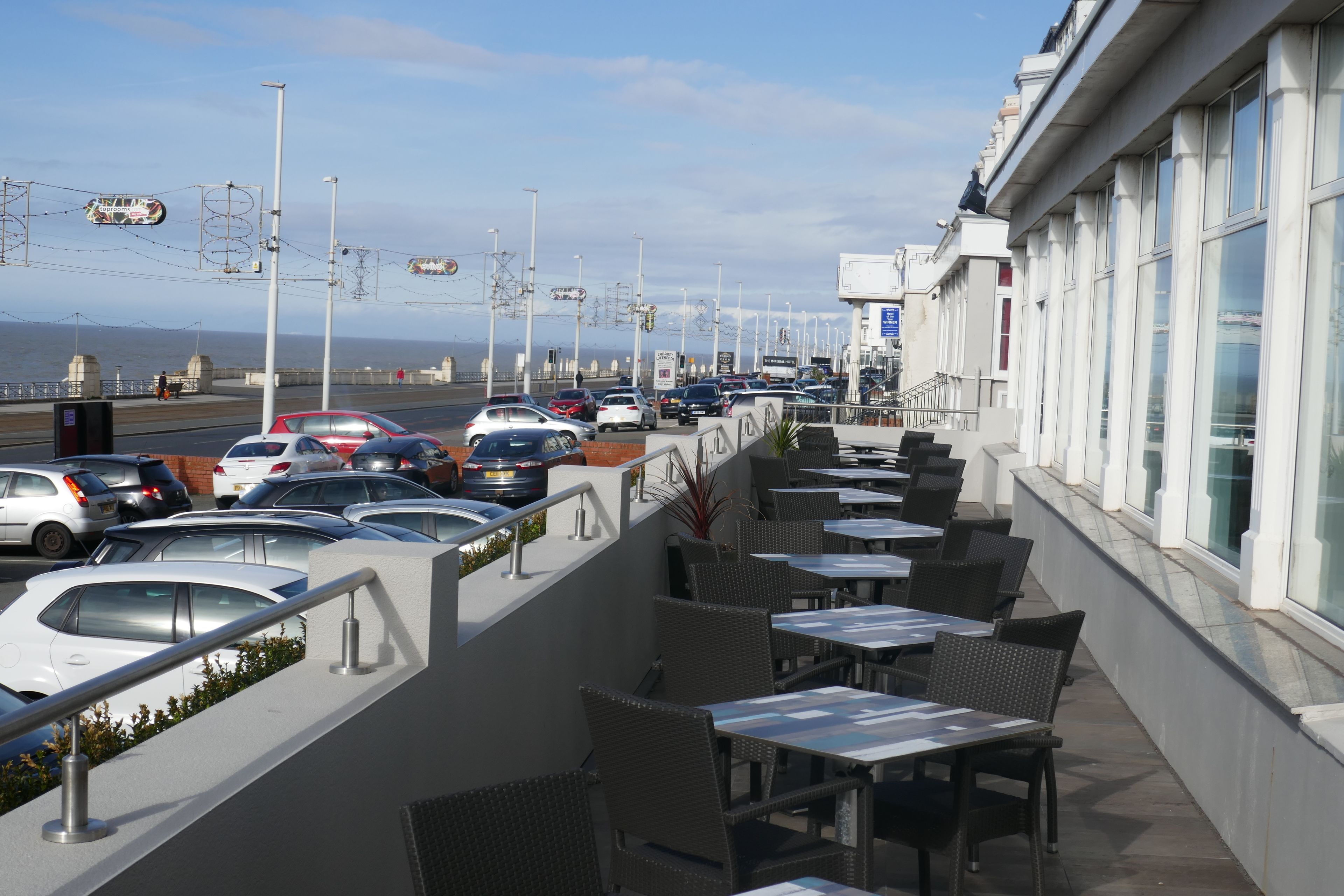 A seaside restaurant with outdoor seating overlooking the ocean, a parking lot, and a view of the beach.