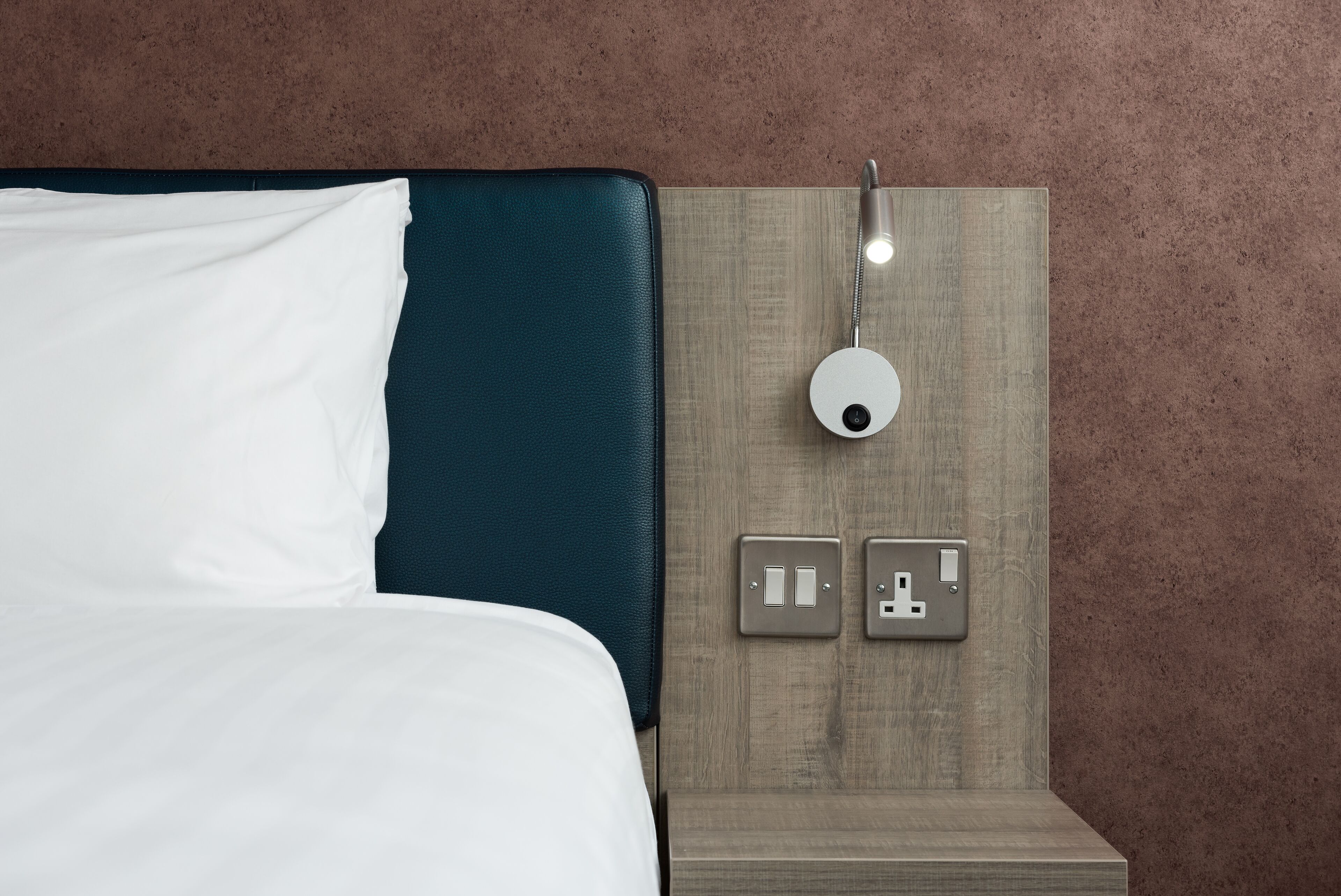A neatly made bed with white linens, a dark blue headboard, and a wall-mounted light fixture with a circular design.
