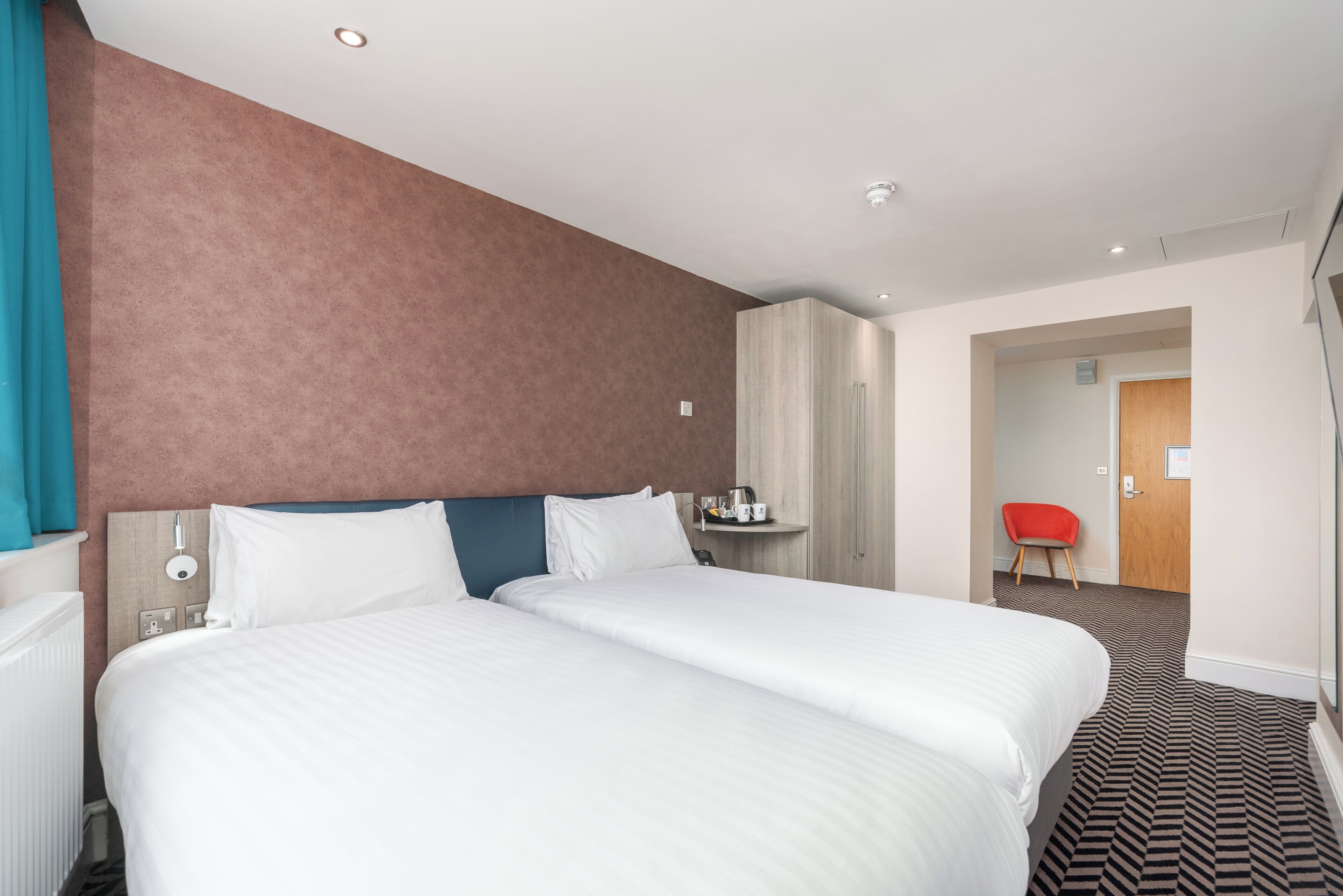 A hotel room with two beds, a red chair, a wooden wardrobe, and a small table with a kettle.