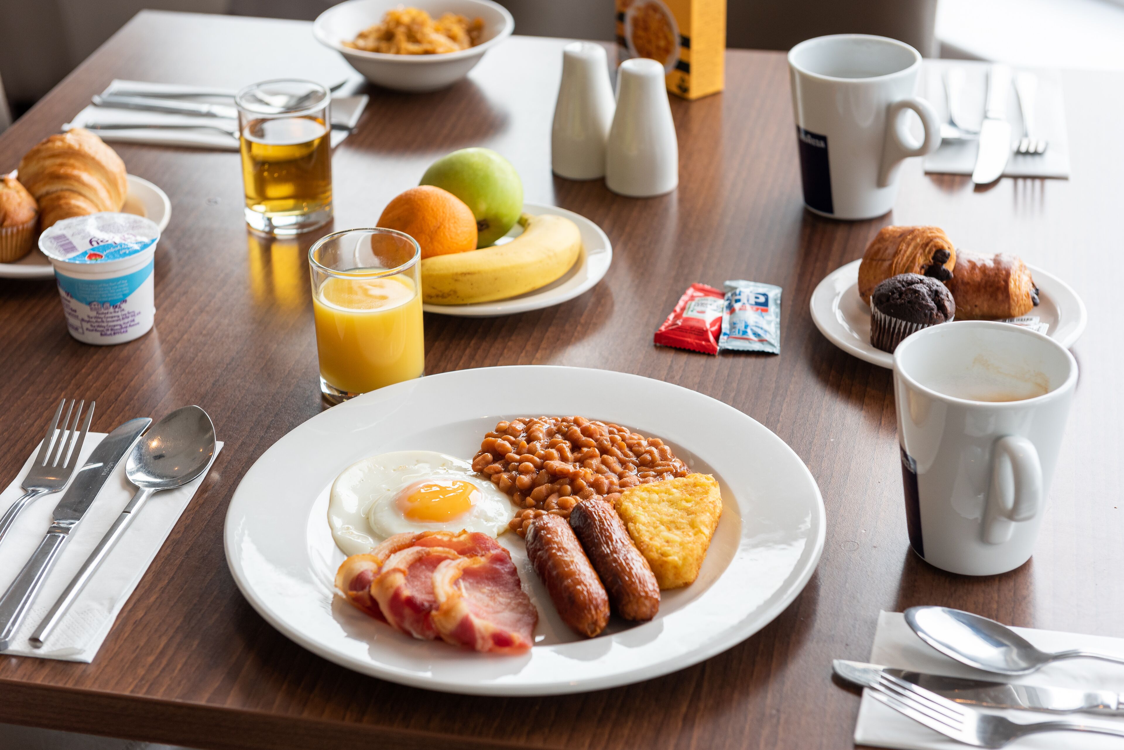 Una mesa de desayuno con un plato de huevos, frijoles, salchichas y tostadas, un vaso de jugo de naranja y una taza de café.