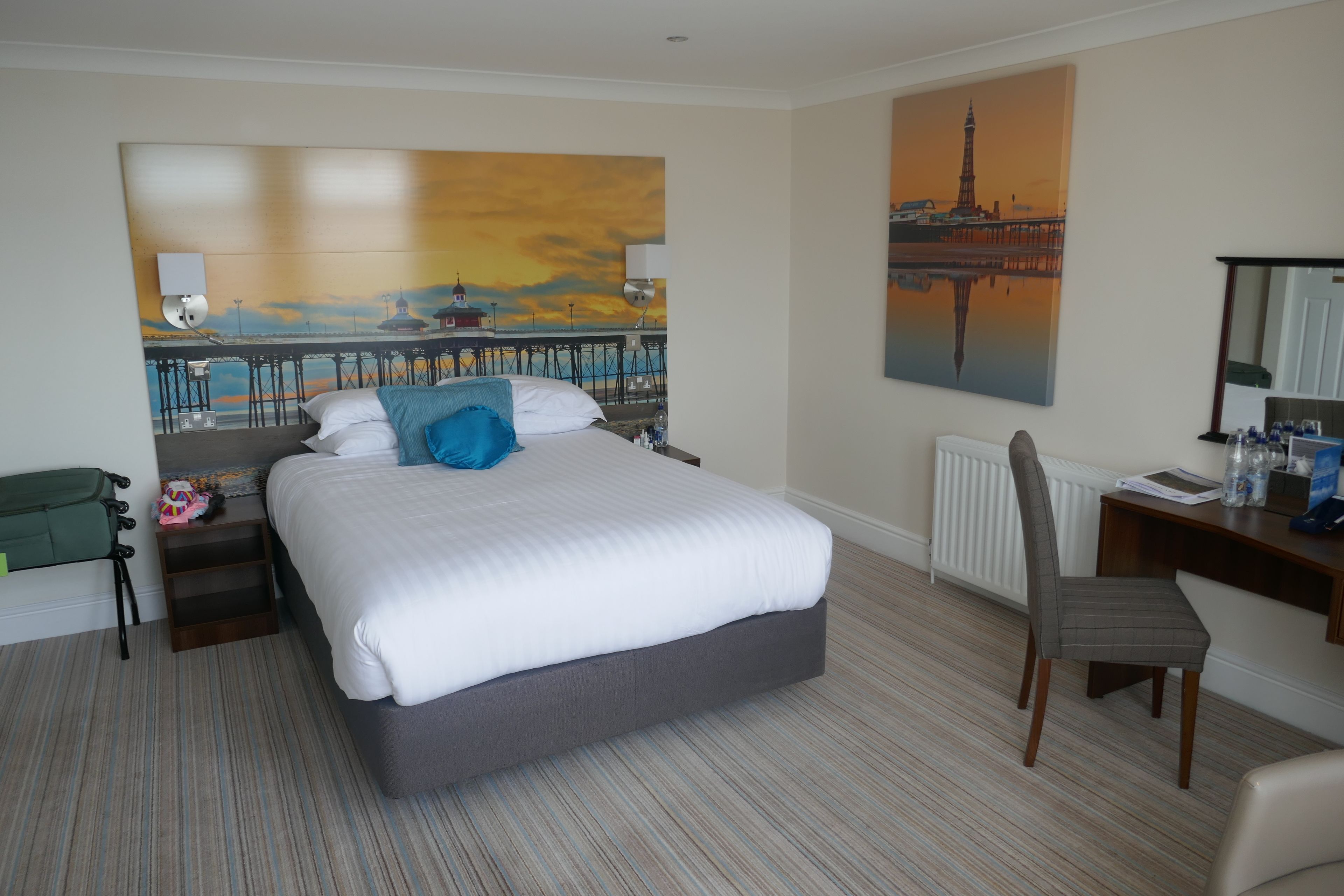 A hotel room with a large bed, a desk, a chair, a radiator, and artwork on the walls.
