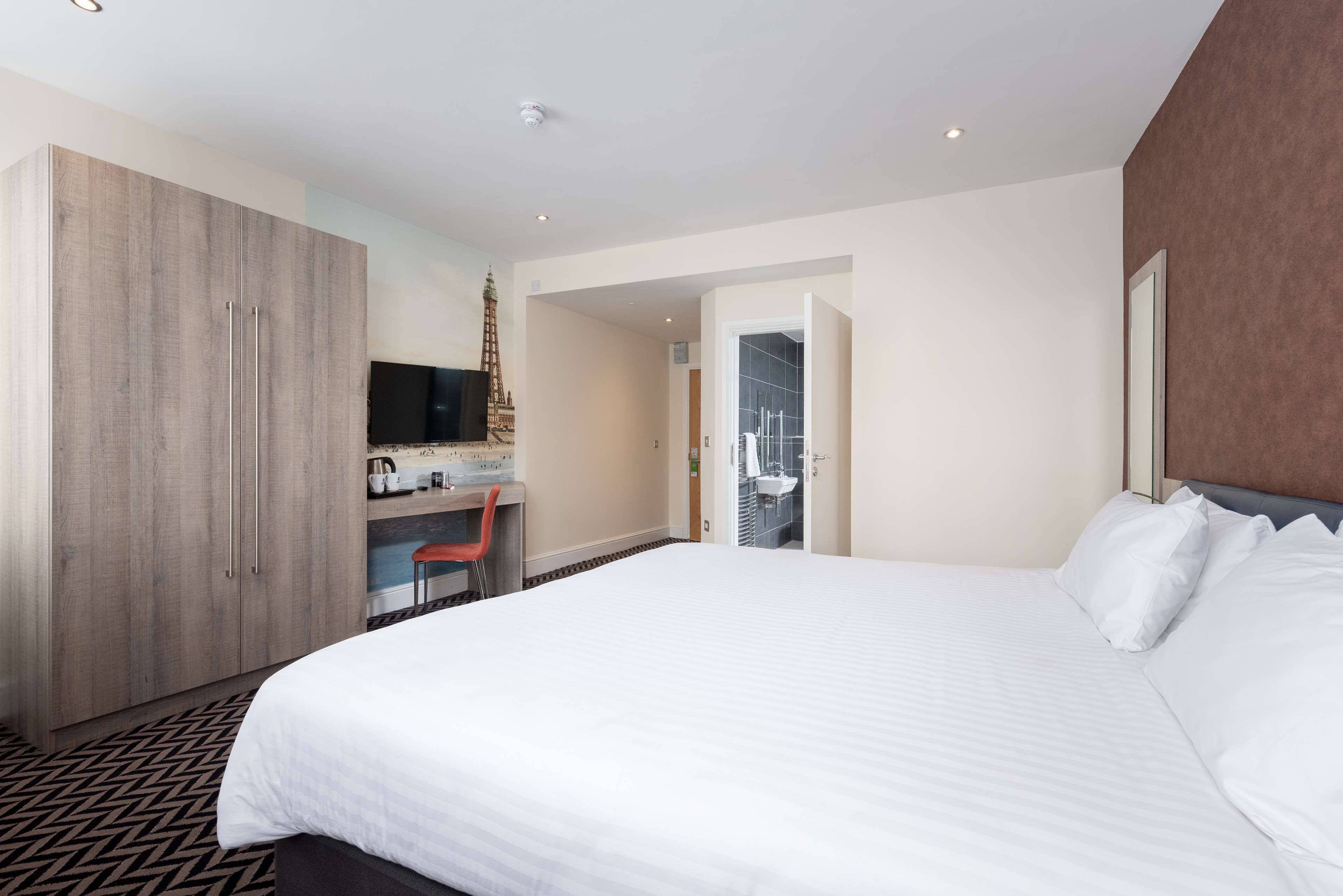 A hotel room with a large bed, a wooden wardrobe, a desk with a chair, a television, and a view of the bathroom.