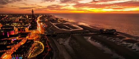 An aerial view of a coastal city at sunset with a prominent tower, a well-lit waterfront promenade, and a beach with visible sand patterns.