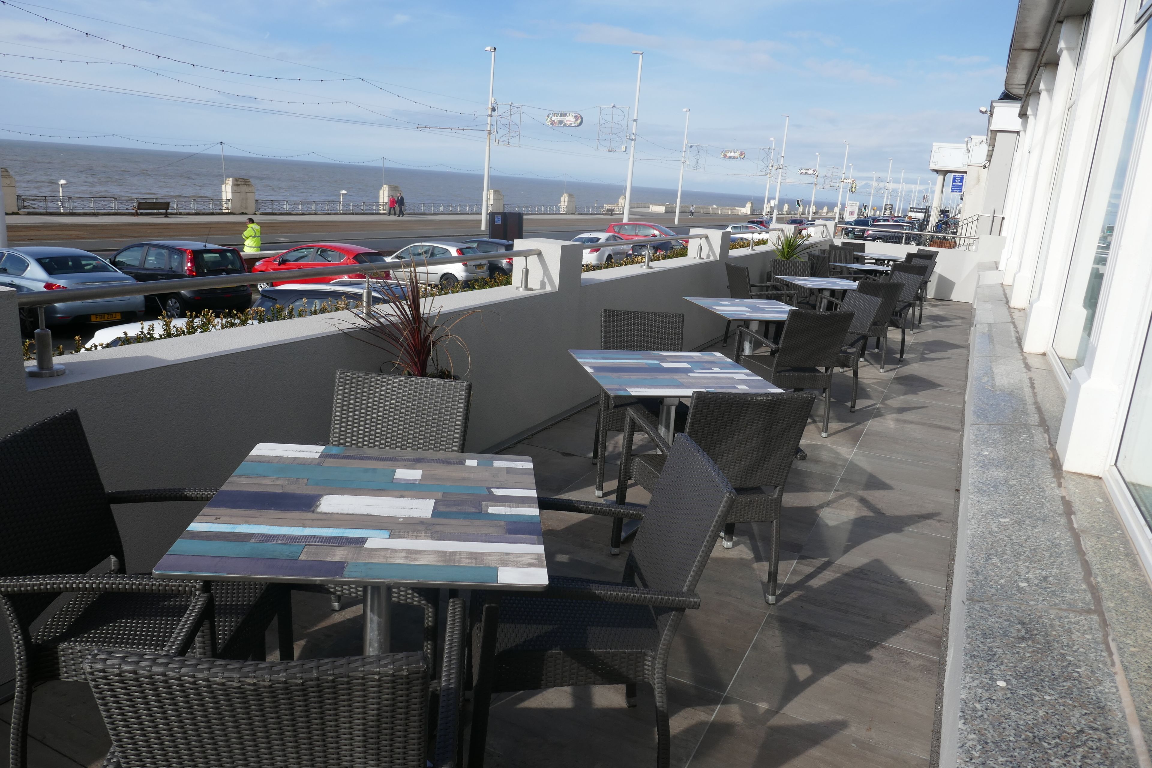Een terras met tafels en stoelen, met uitzicht op een parkeerplaats met auto's en de zee.
