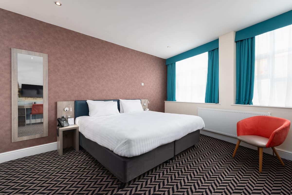 A modern hotel room with a large bed, a desk, a chair, and a mirror.