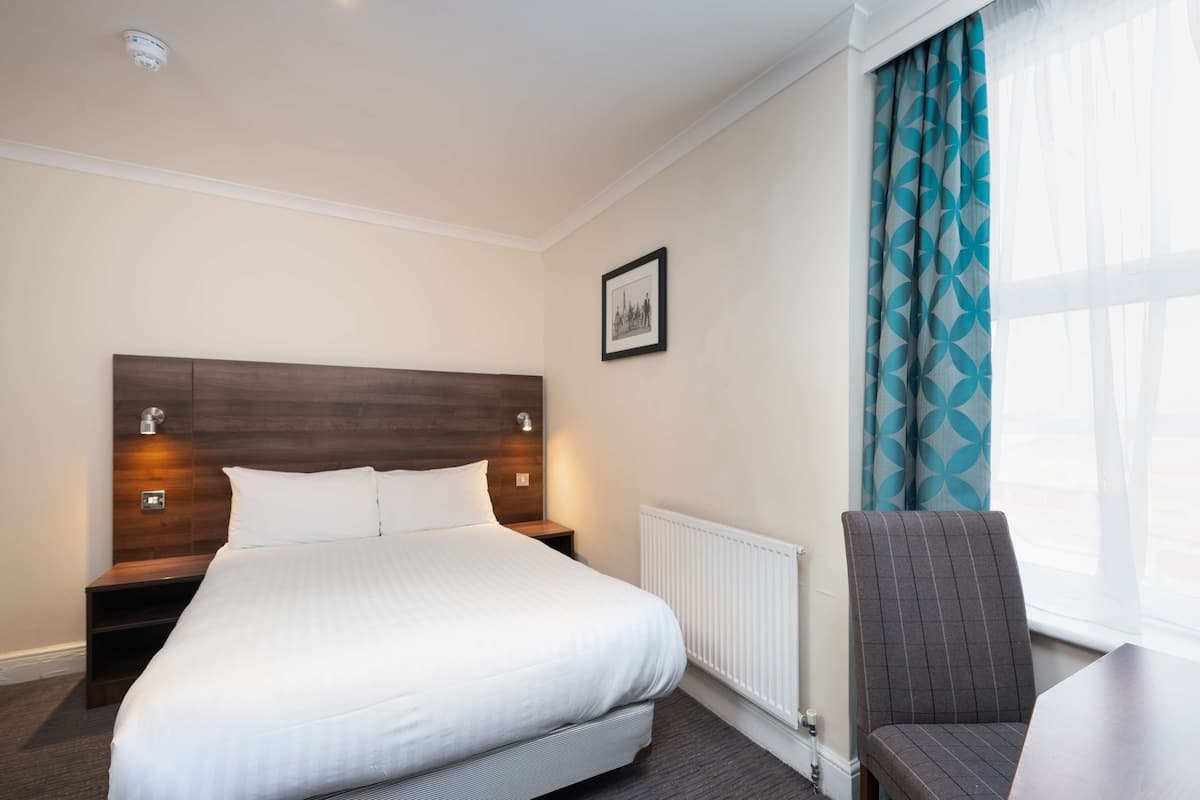 A hotel room with a large bed, a nightstand, a chair, a framed picture on the wall, and a window with curtains.