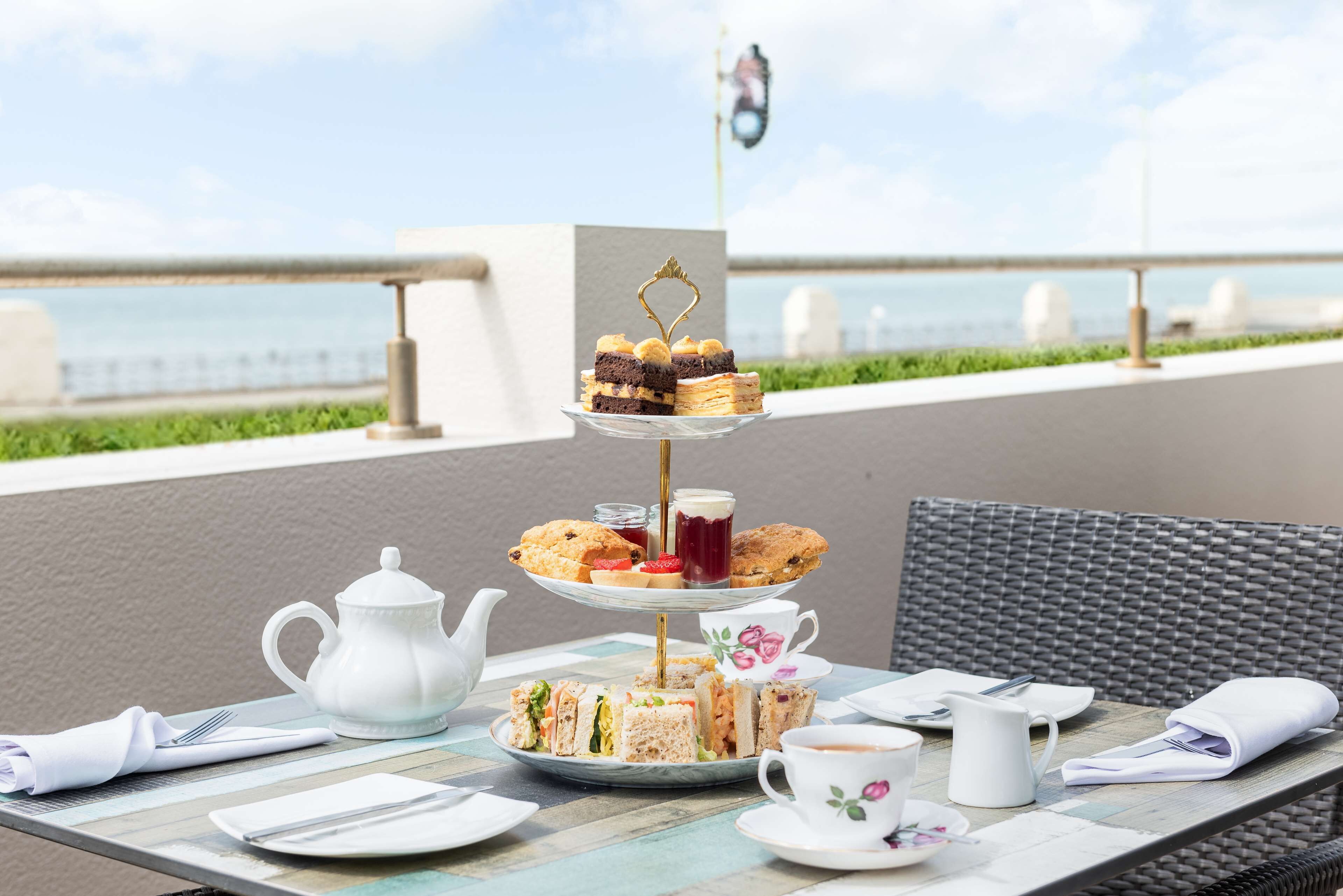 A three-tiered dessert stand with various pastries and sandwiches, a white teapot, cups, and a wicker chair on an outdoor table with a view of the sea.