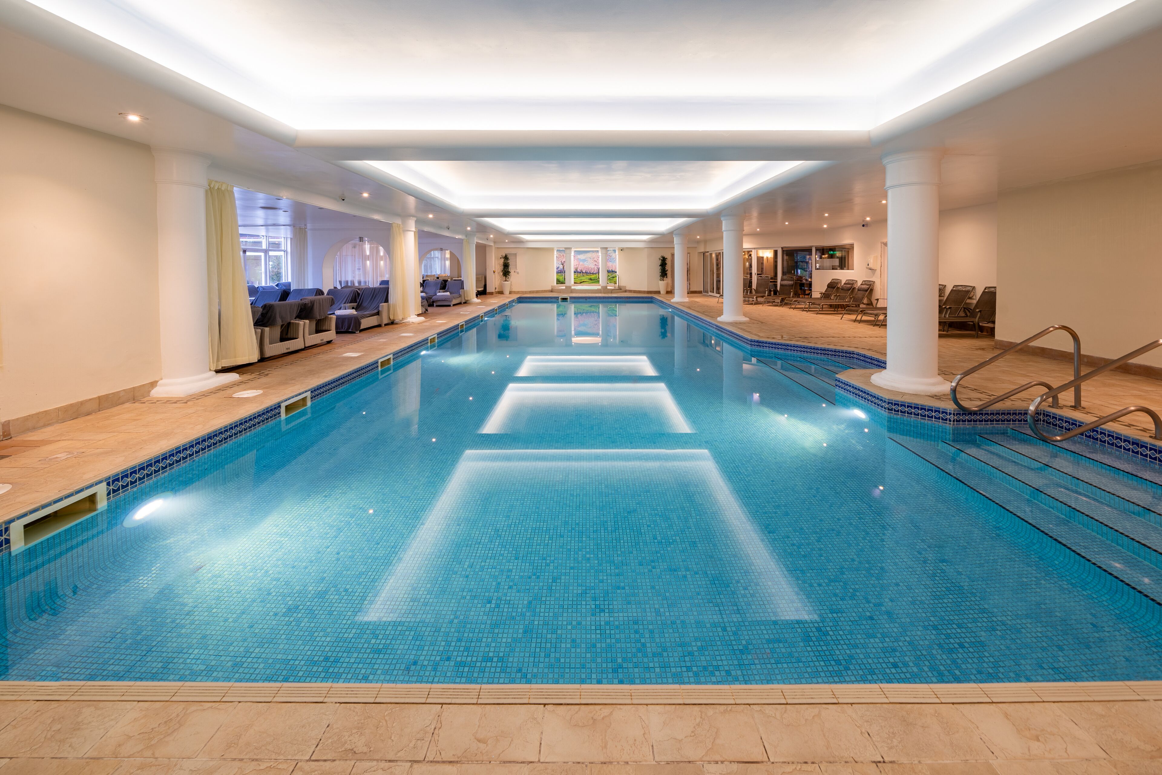 A large indoor swimming pool with clear water, surrounded by lounge chairs and a tiled floor.