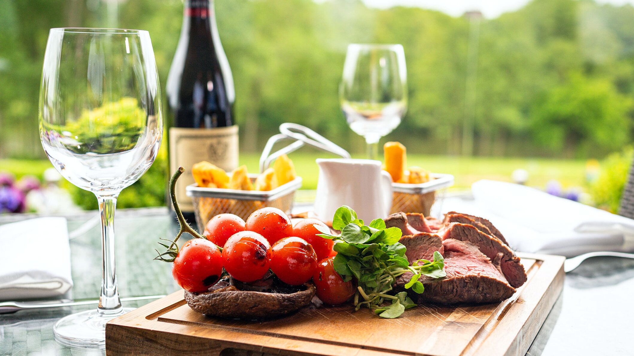 A wooden cutting board with sliced steak, cherry tomatoes, and greens, accompanied by two glasses of wine and a bottle of wine.