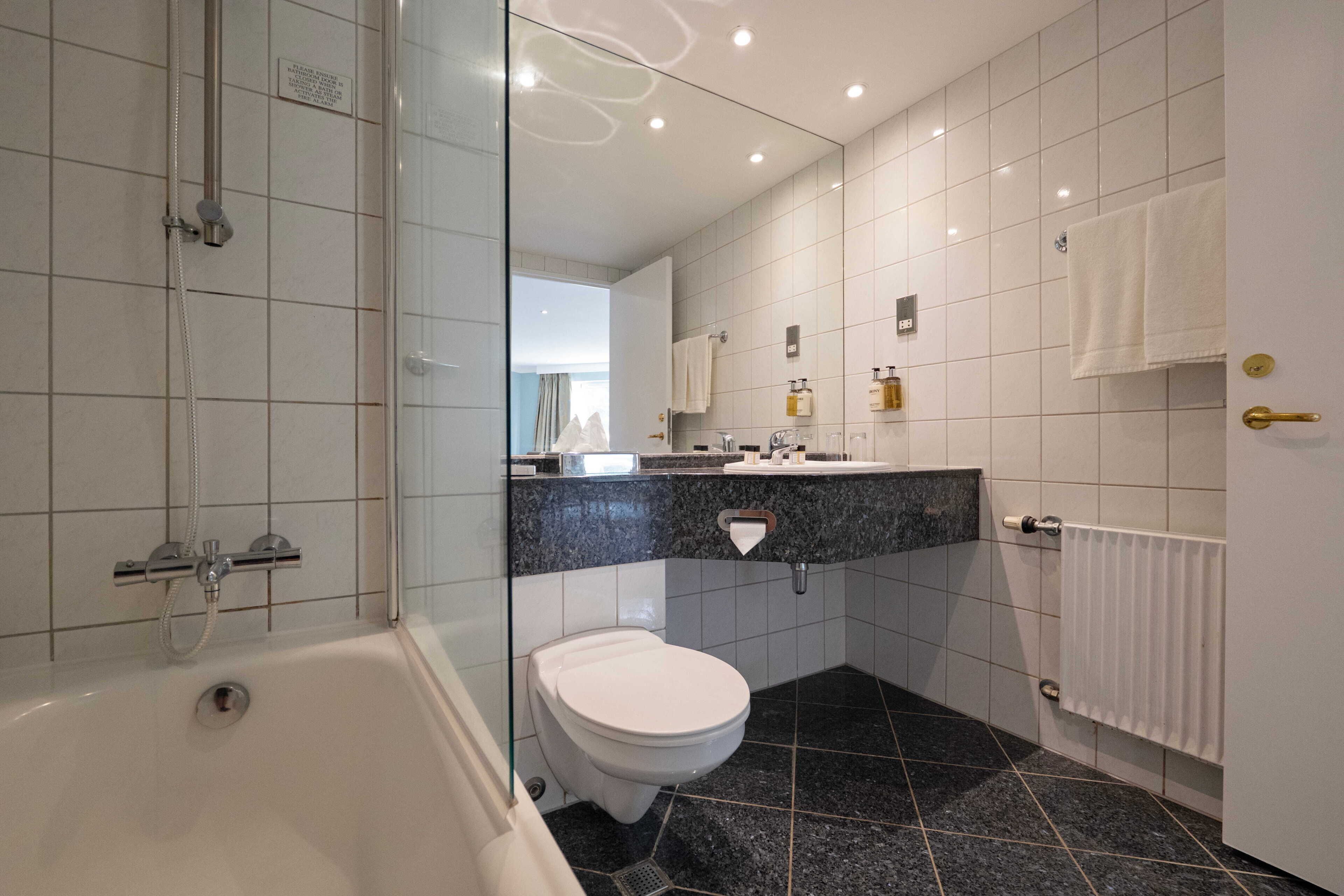 A modern bathroom with a glass-enclosed shower, a white toilet, a marble-tiled floor, and a vanity with a mirror and toiletries.