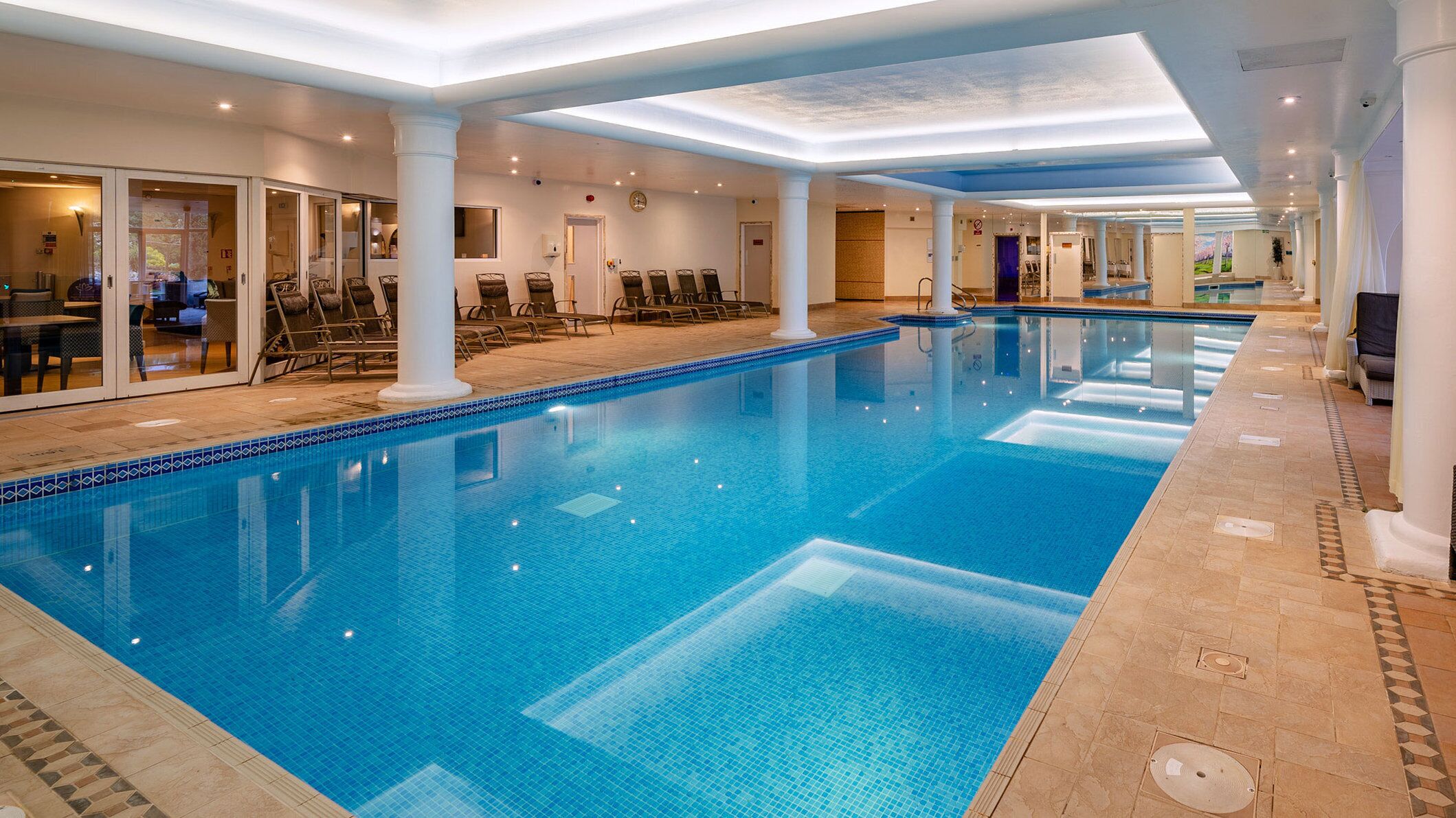 Une grande piscine intérieure à l’eau d’un bleu limpide, entourée de chaises longues et d’un espace de repos abrité.