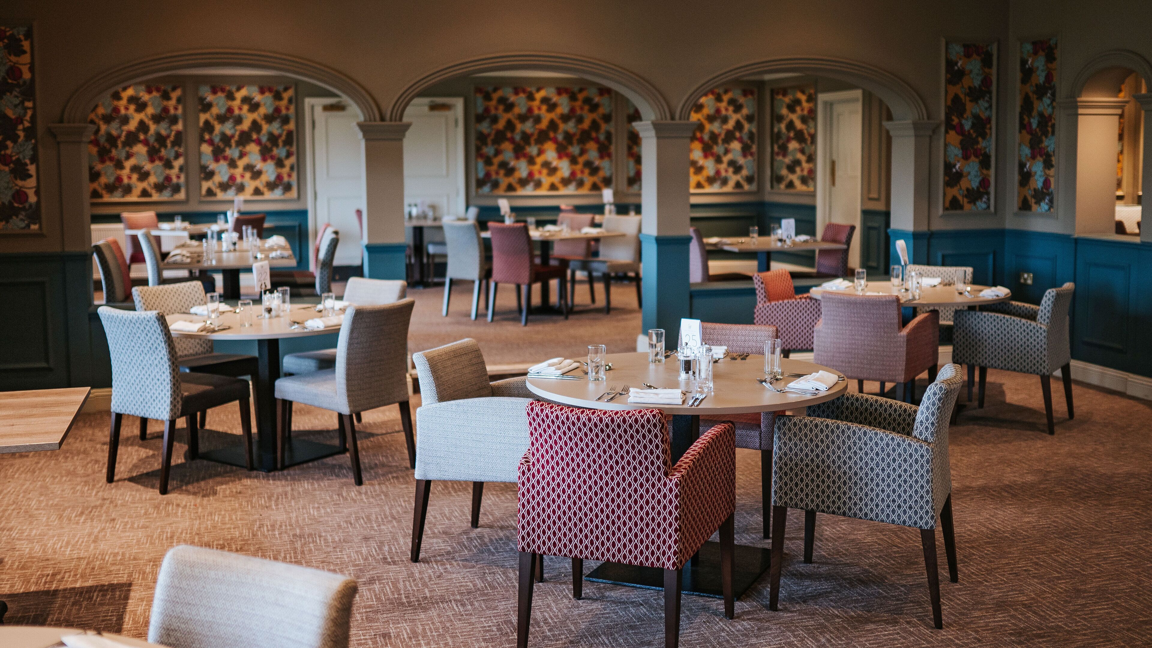 Ein Restaurant mit mehreren Tischen und Stühlen, das eine strukturierte Wand und Rundbogenpfeiler zeigt.