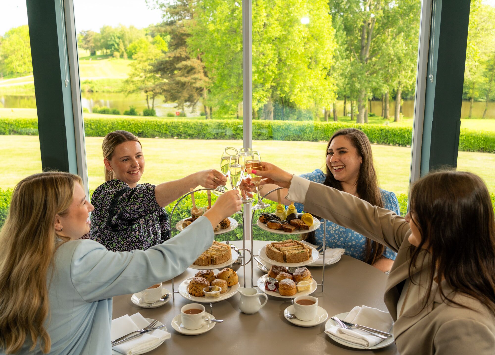 A group of people toasting at a table with a variety of pastries and coffee, set in a room with large windows overlooking a garden.