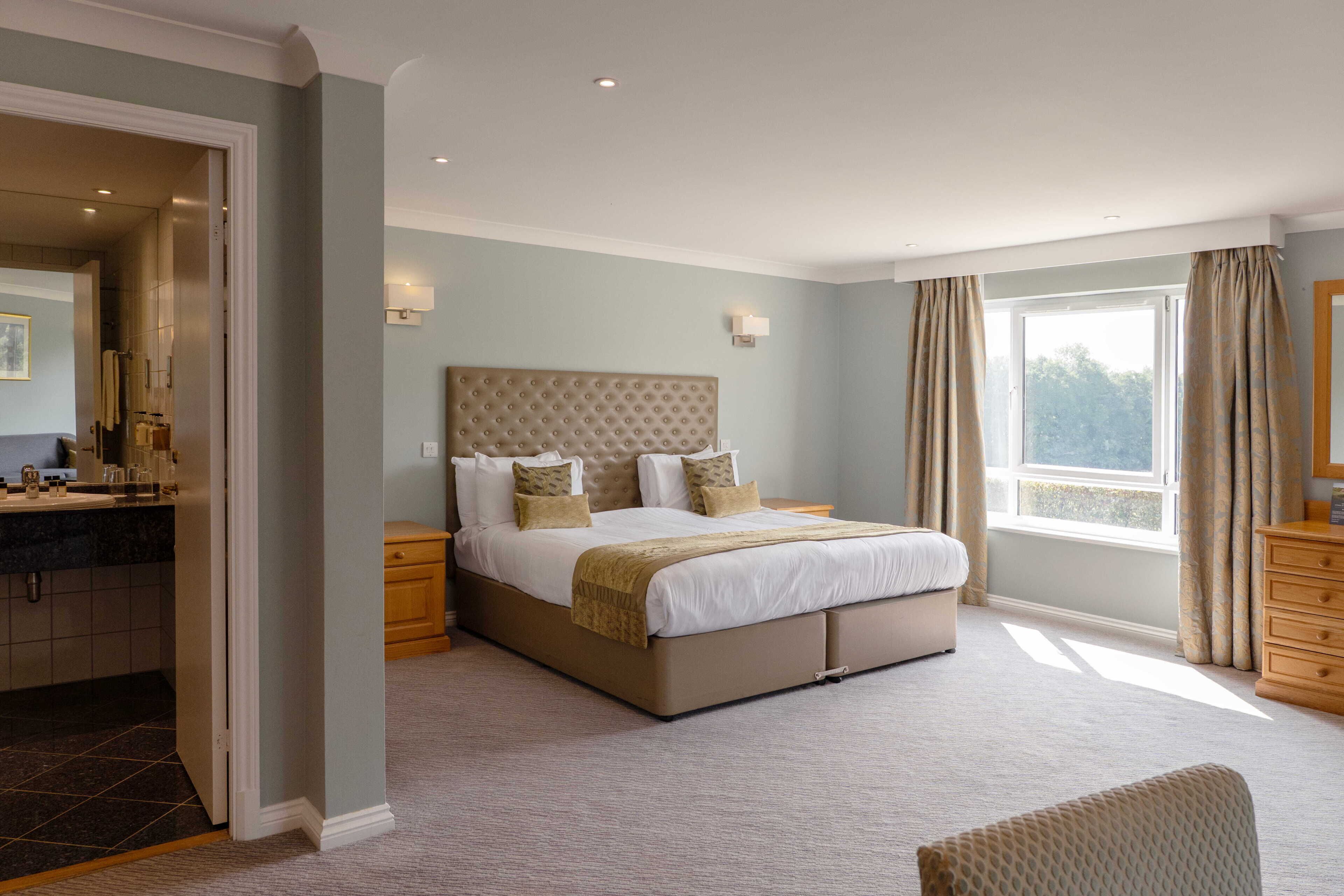 A bedroom with a large bed, a bathroom with a mirror, and a window with curtains.