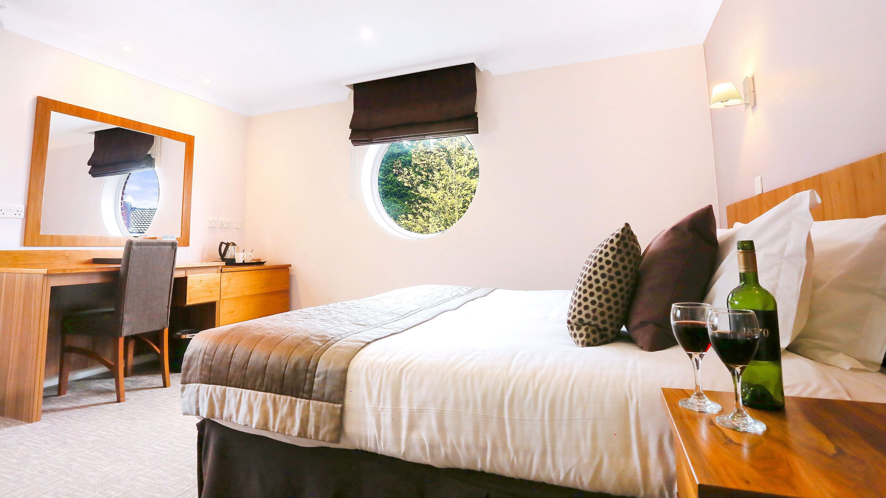 A hotel room with a bed, a desk with a chair, a mirror, a window with a view of trees, and a nightstand with a bottle of wine and glasses.