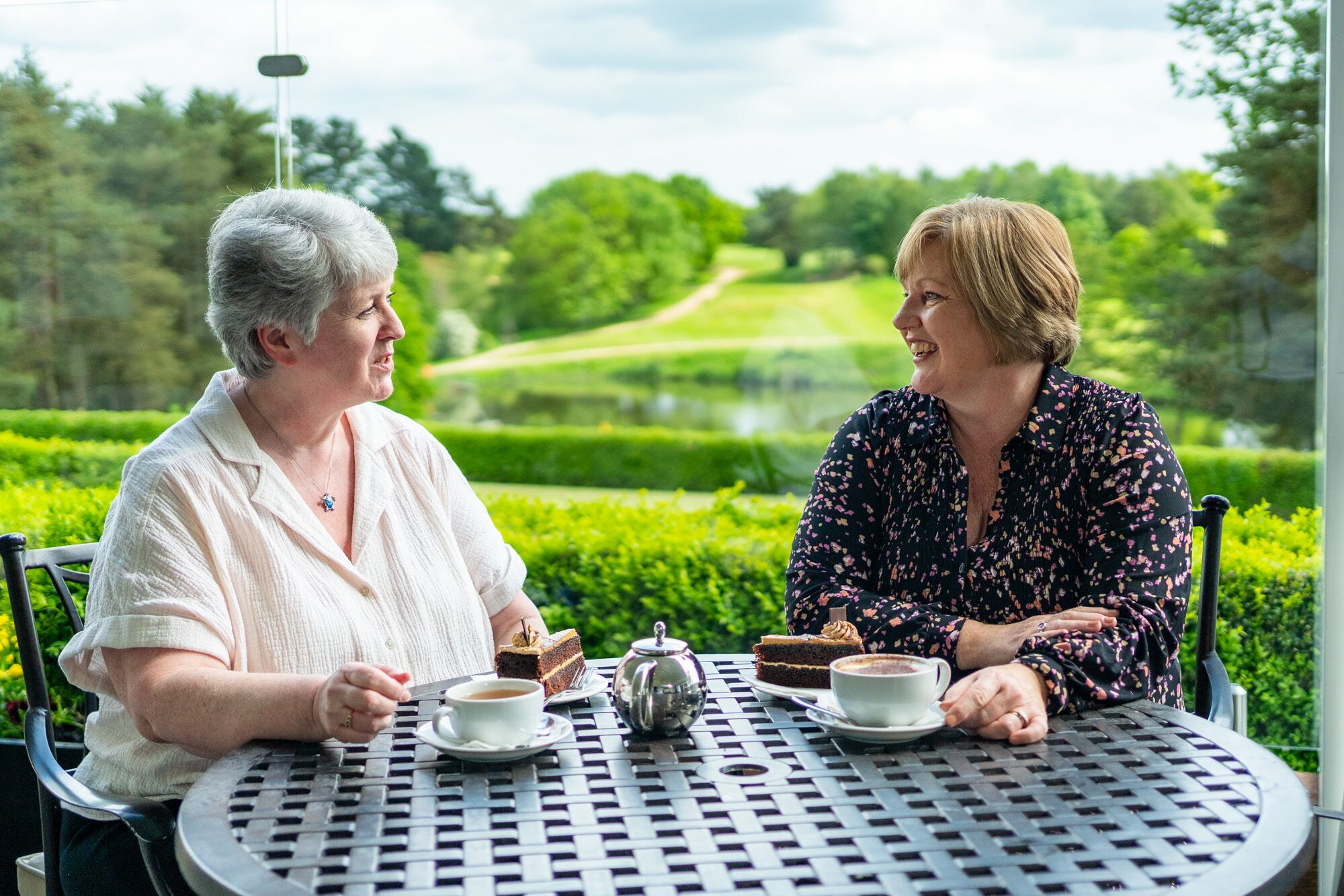 Two people sitting at a round table outdoors with a view of a manicured garden and a pathway.