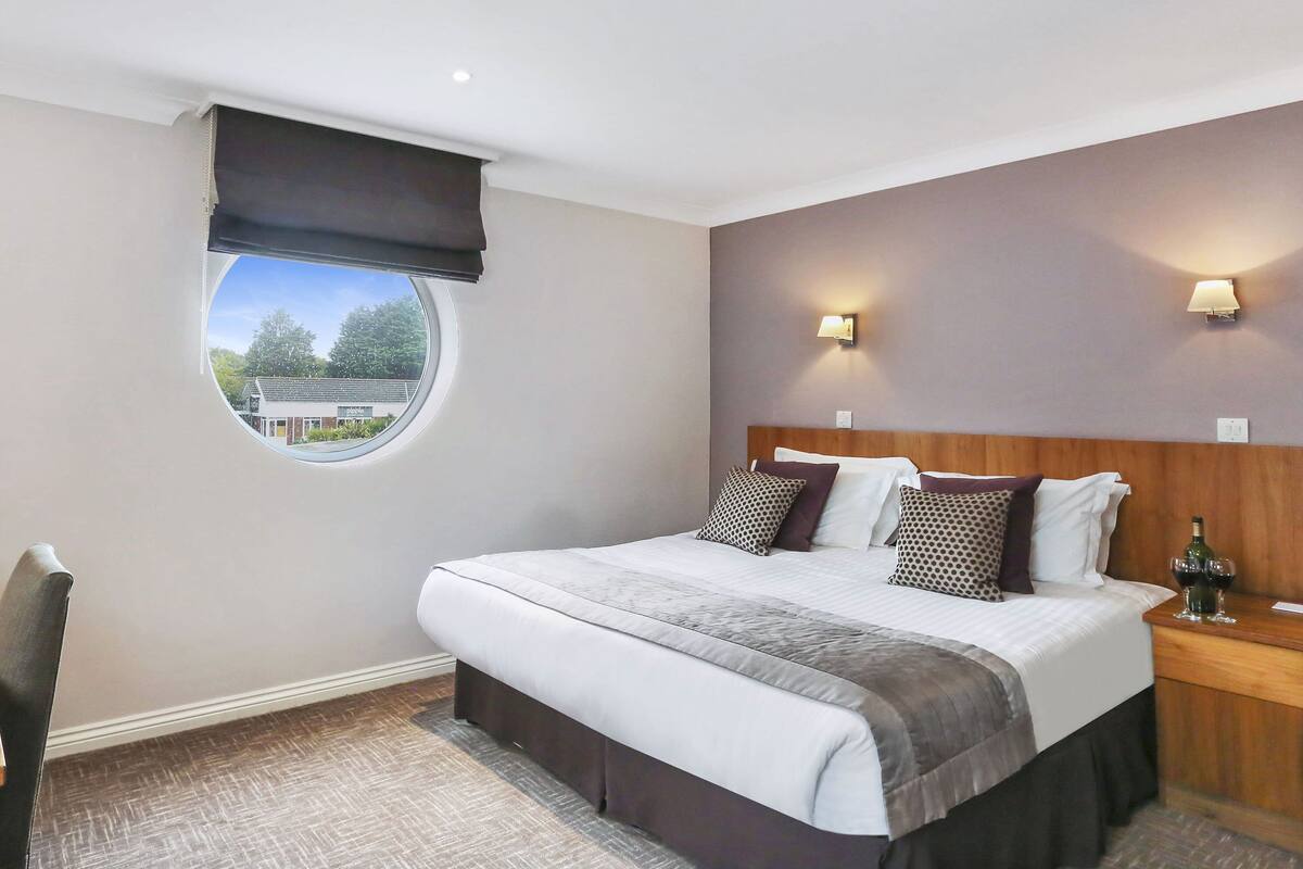 A hotel room with a large bed, a round window, a desk with a coffee maker, and two wall-mounted lamps.