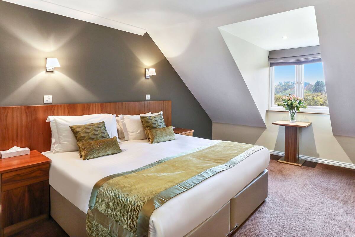 A bedroom with a sloped ceiling, a bed with white linens, a wooden headboard, two bedside tables, a nightstand, a small table with a vase, and a window with a view.