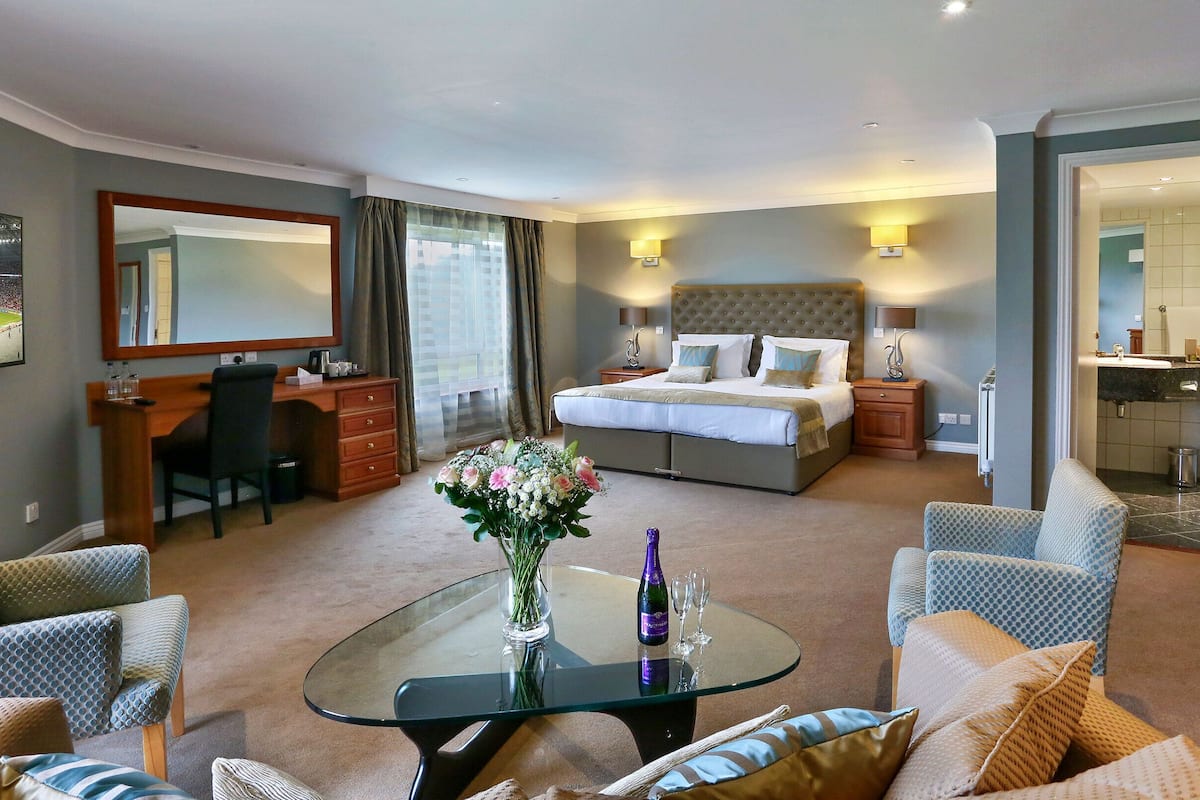 A hotel room with a bed, a desk, a chair, a glass table with a vase and champagne, and a view of a sports stadium.