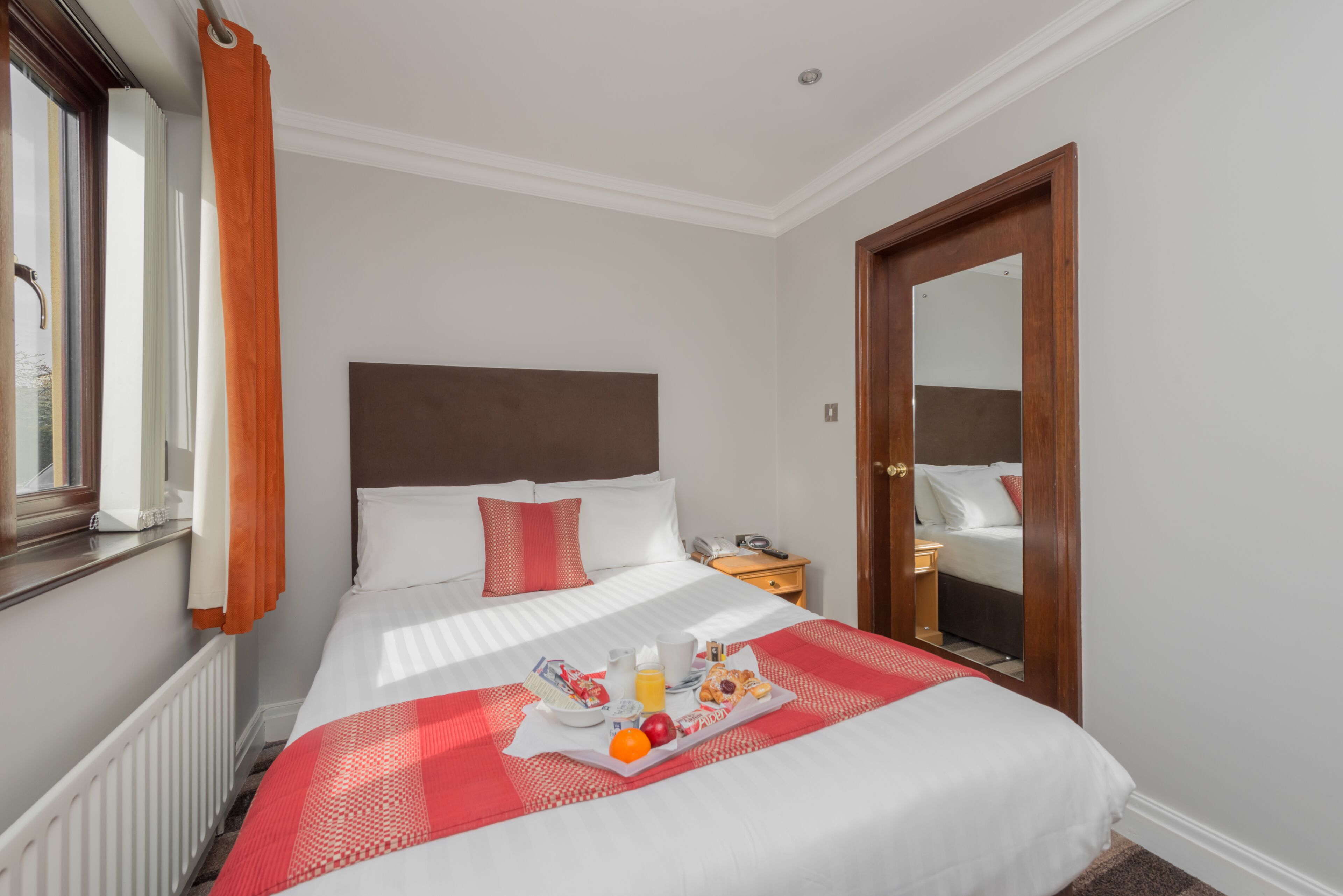 A hotel room with a neatly made bed, a bedside table with a phone, and a tray of breakfast items.