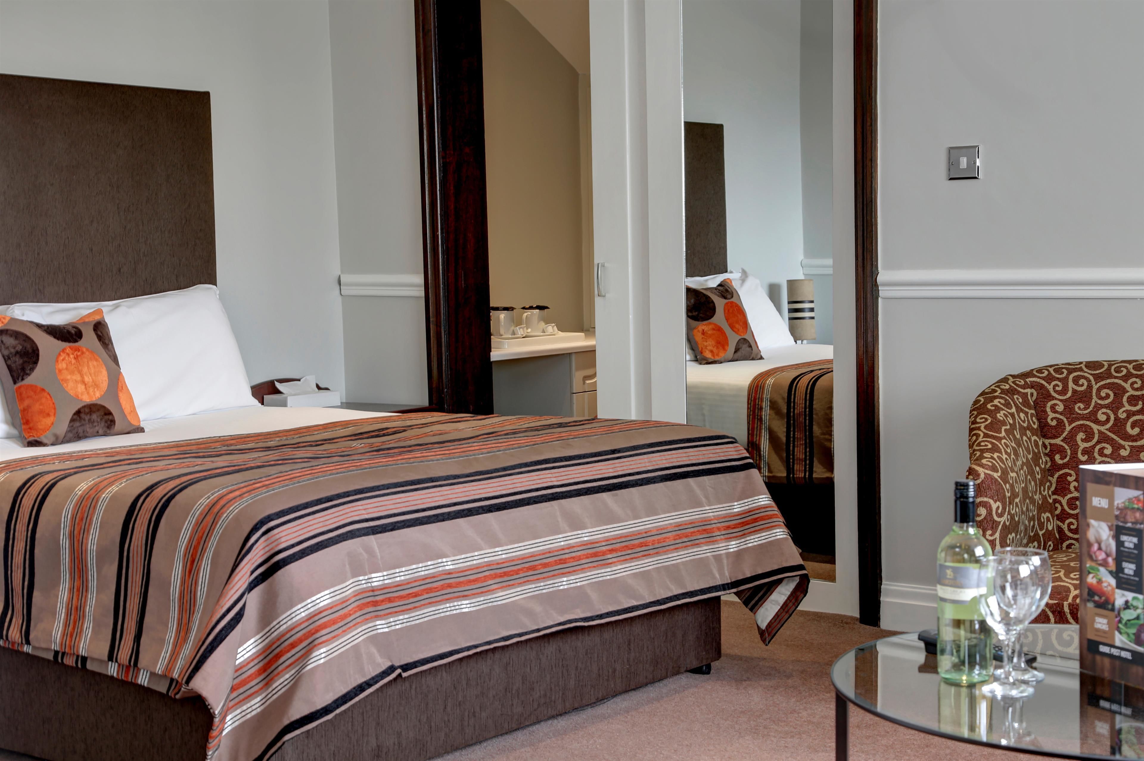 A hotel room with a large bed, a bedside table with a bottle and glass, and a chair with a patterned cushion.