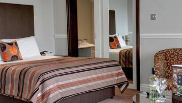 A hotel room with a large bed, a bedside table with a bottle and glass, and a chair with a patterned cushion.