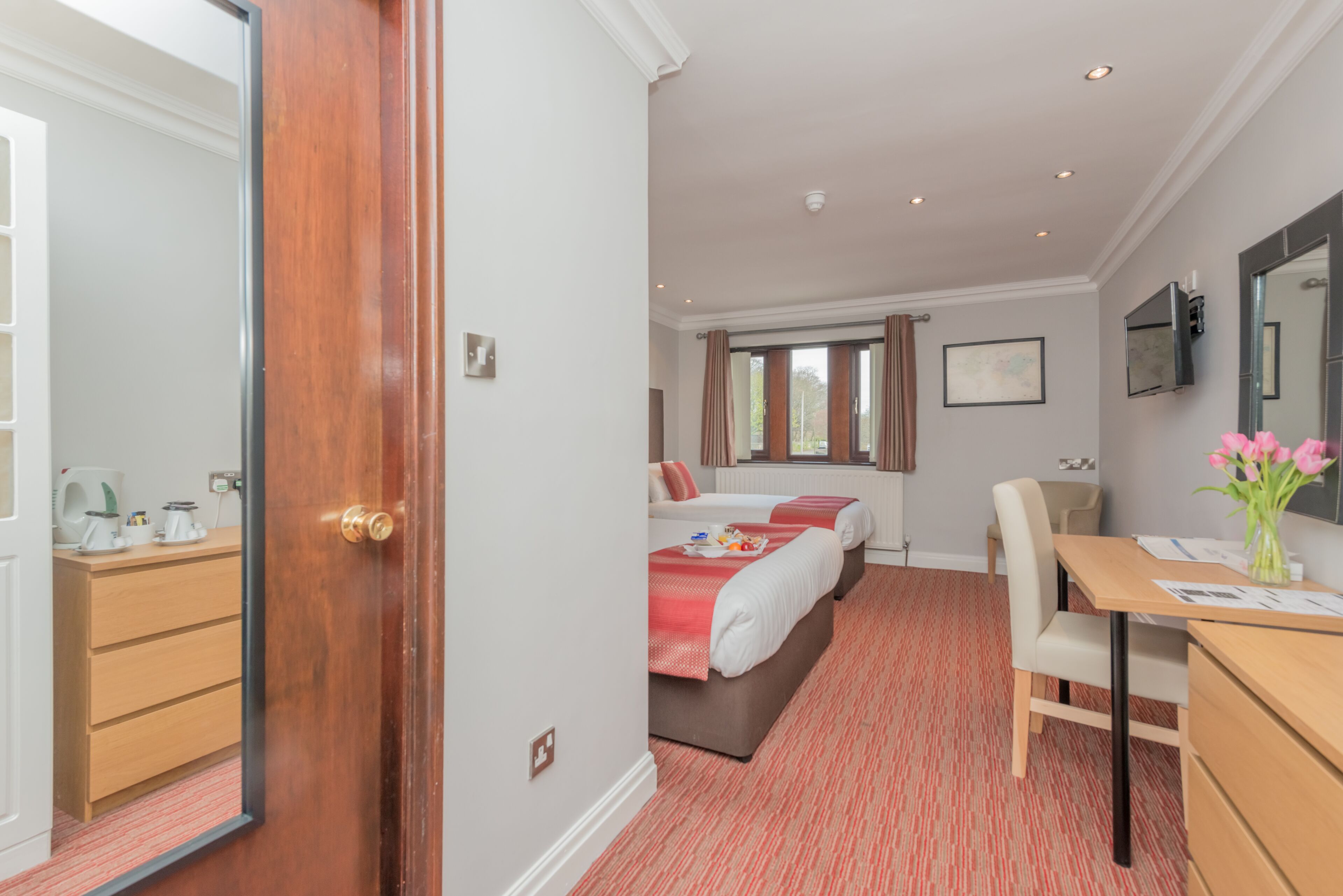 A hotel room with a wooden door, a desk with a chair, two beds, a TV, and a window with curtains.