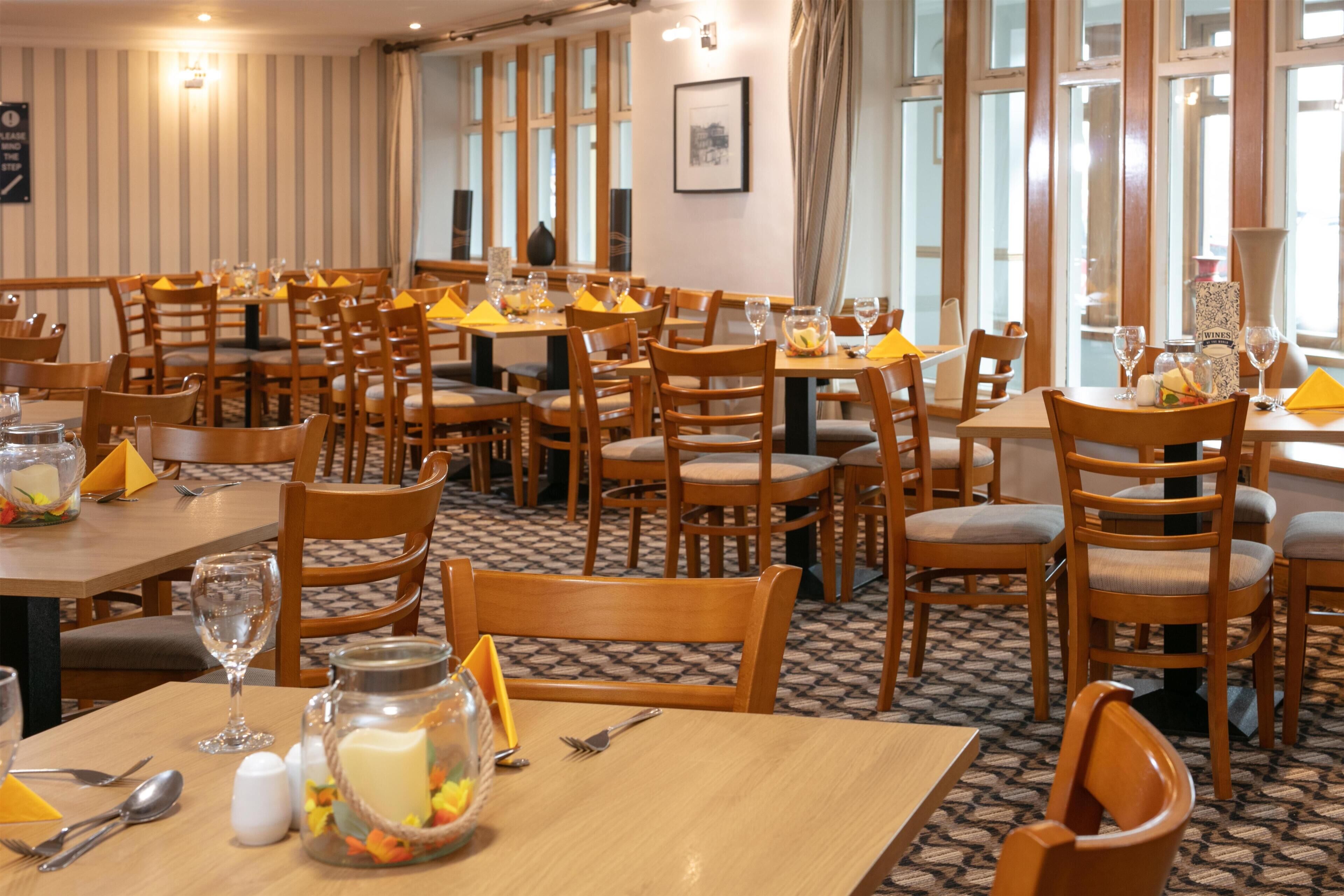 A dining area with multiple tables and chairs, arranged in a spacious layout with large windows allowing natural light.