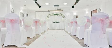 Un salón de bodas con un pasillo blanco, decoración floral y sillas blancas adornadas con cintas rosas.