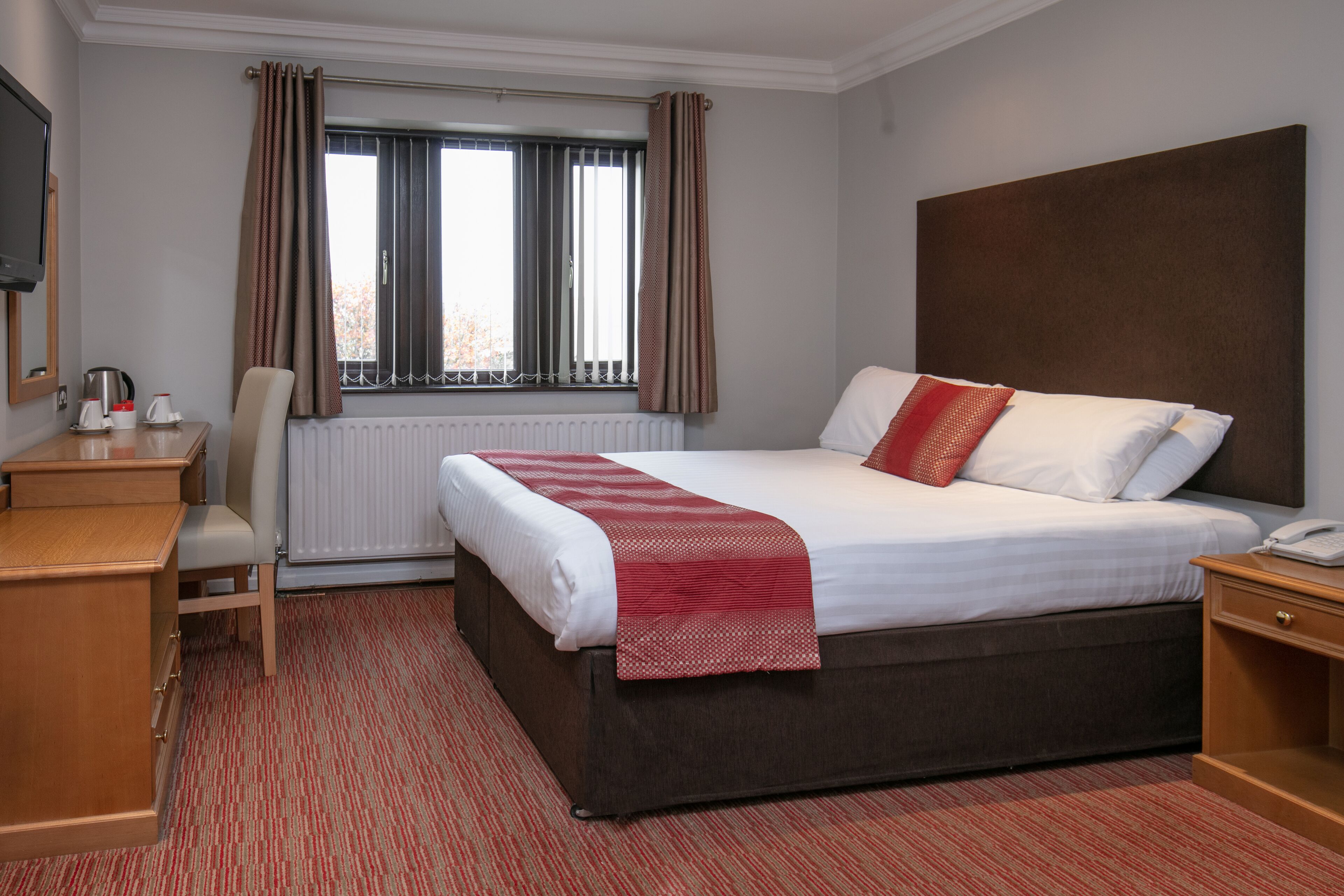 A hotel room with a bed, a desk, a chair, a radiator, and a window with curtains.