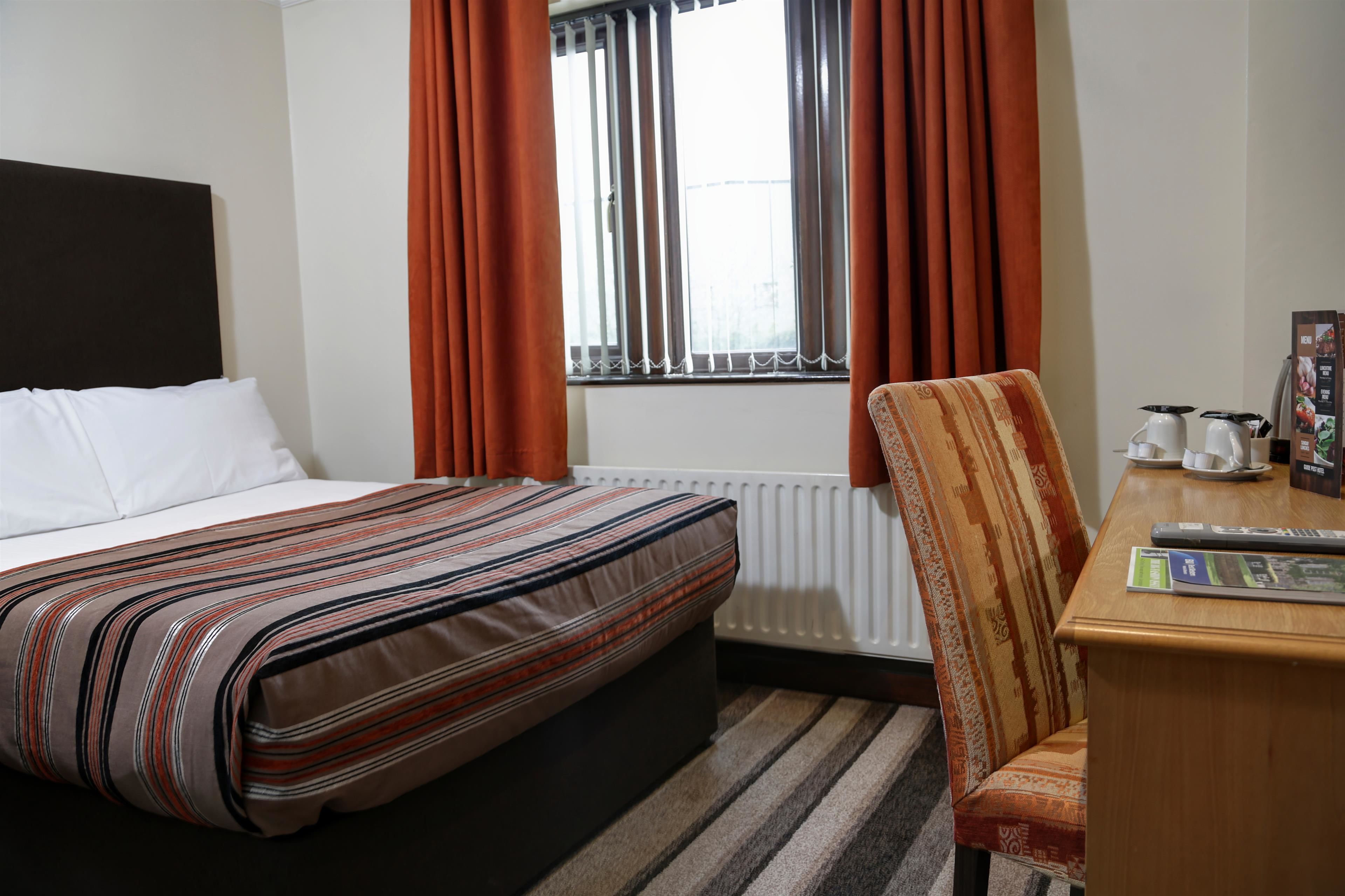 Habitación de hotel con una cama, una silla, una mesa con hervidor y tazas, y un ventanal con cortinas.