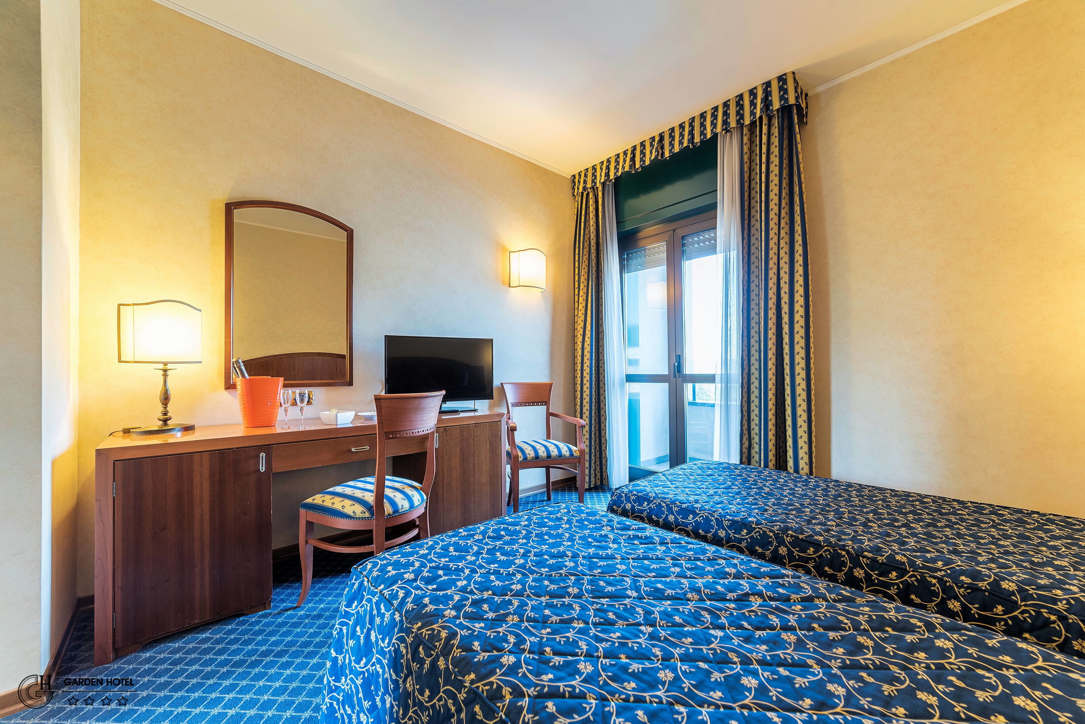 Una habitación de hotel con escritorio, dos sillas, televisor y dos camas con ropa de cama estampada.