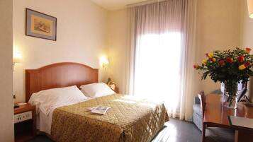 Ein Hotelzimmer mit Bett, Schreibtisch, Stuhl, Lampe, einem Bild an der Wand und einem Fenster mit Vorhängen.