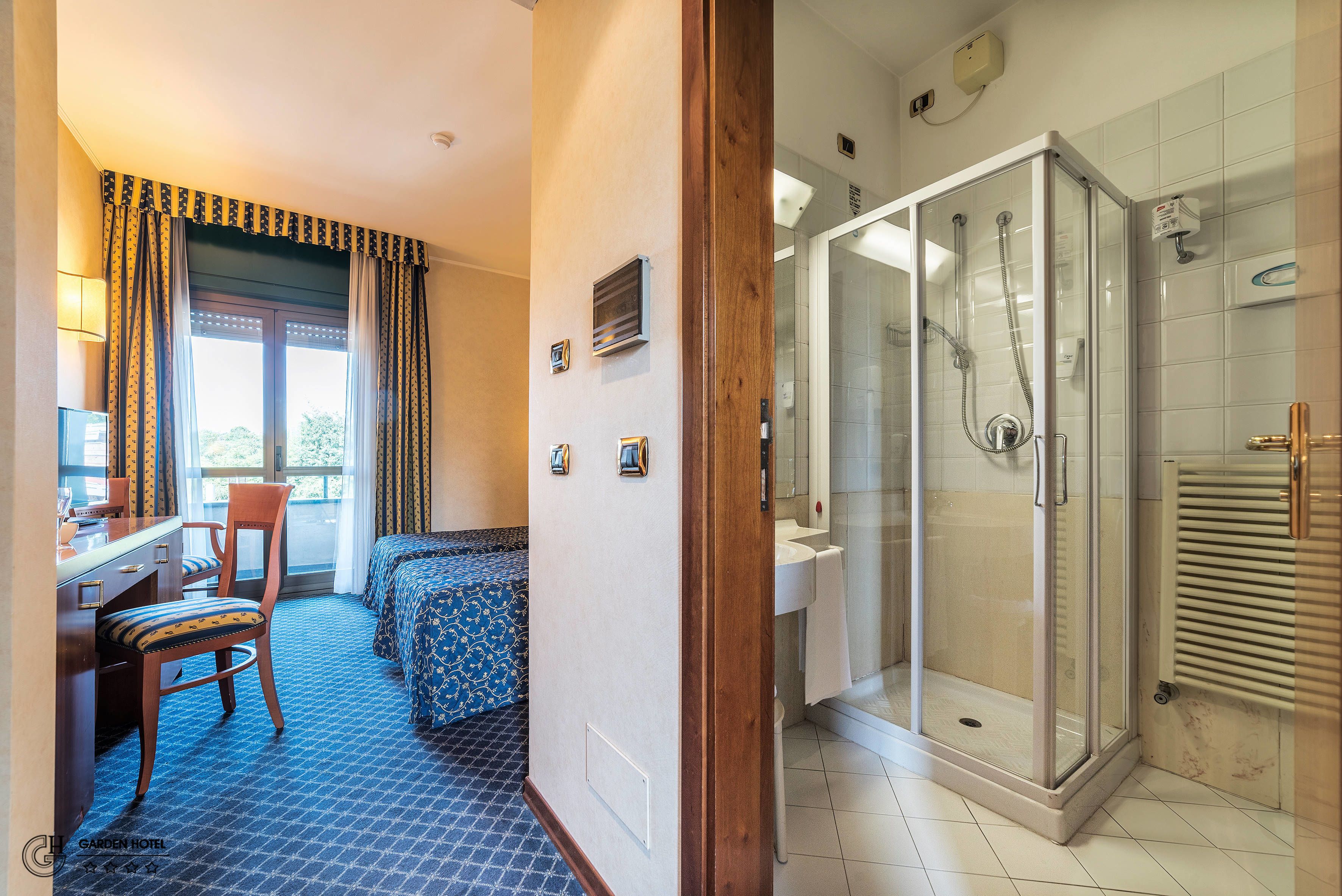 A hotel room with a desk, chair, bed, and a glass-enclosed shower area.