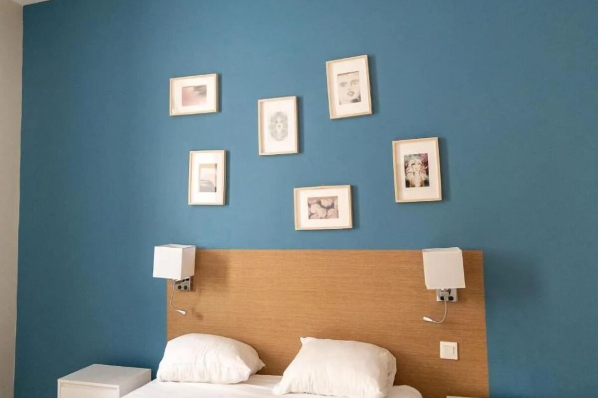 A hotel room with a blue wall, a wooden headboard, and a bed with white bedding. There are six framed pictures on the wall.