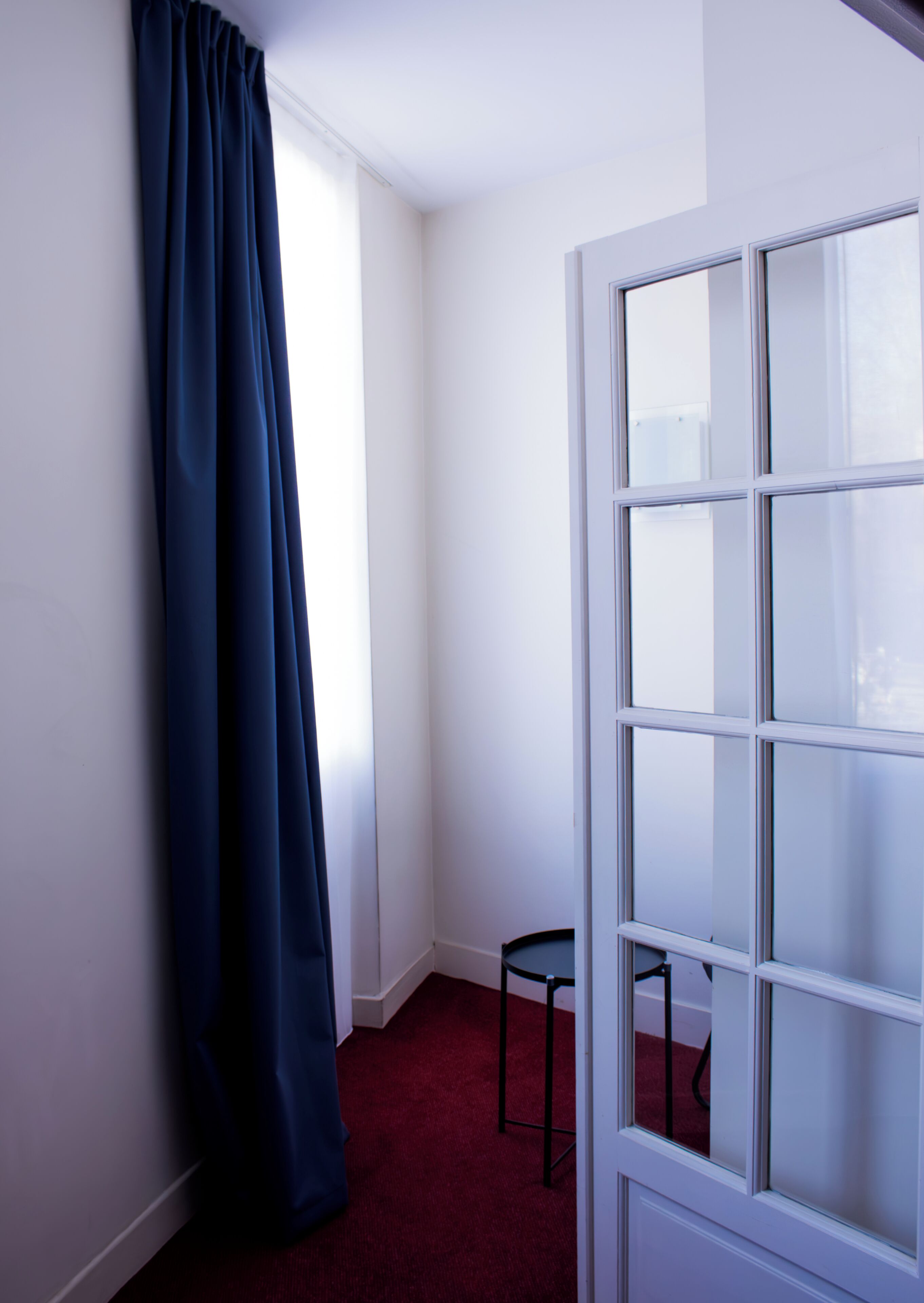Una habitación con alfombra roja, cortina azul oscuro, una pequeña silla negra y una puerta blanca con paneles de vidrio esmerilado.