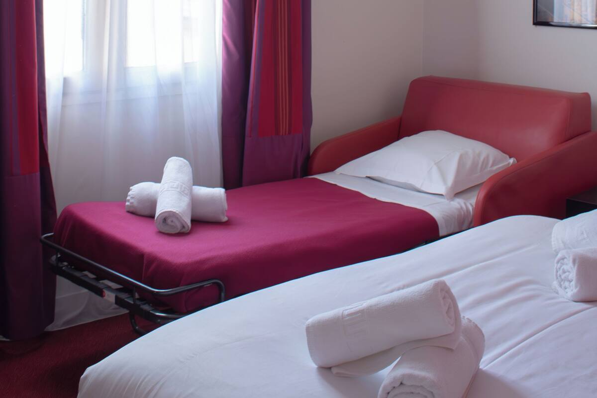 Een hotelkamer met twee bedden, een rode stoel en een raam met dunne gordijnen.