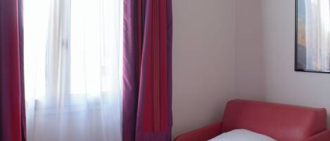 A hotel room with two beds, a red chair, and a window with sheer curtains.