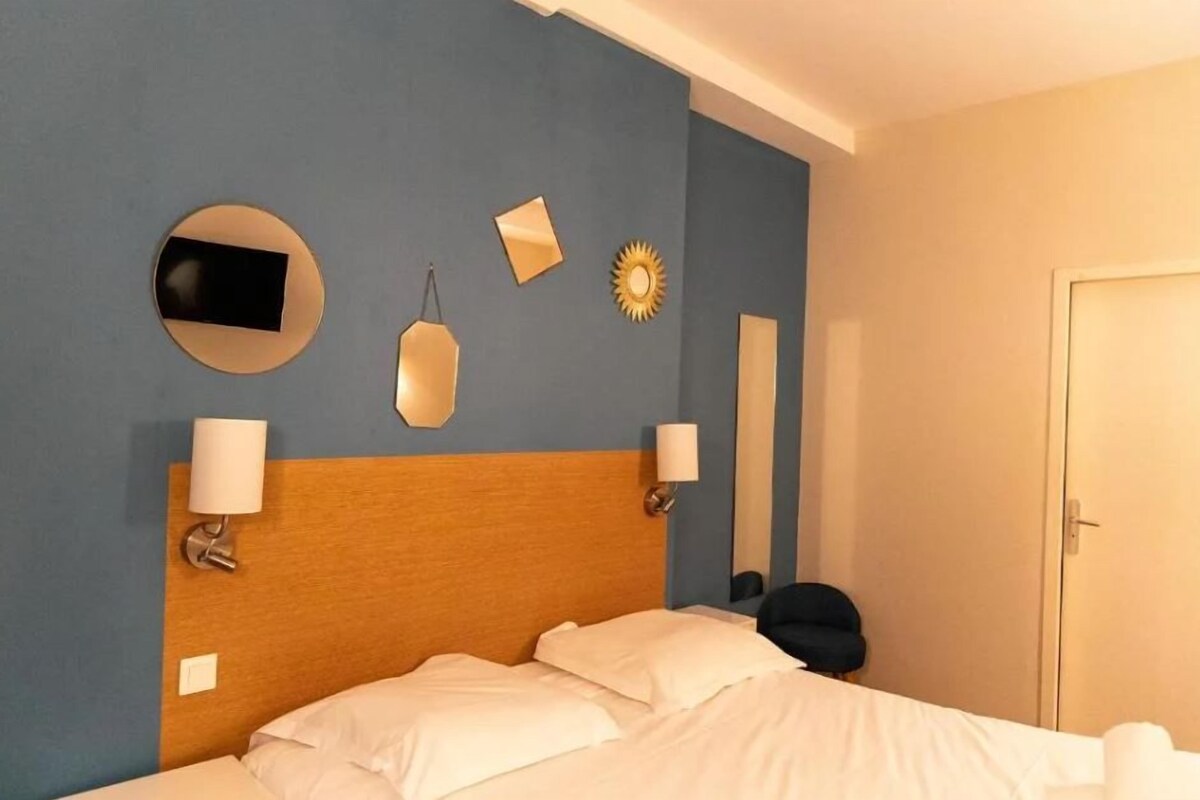 Une chambre d’hôtel comprenant un lit, deux lampes fixées au mur, un miroir rond, un miroir rectangulaire et un élément décoratif en forme de soleil rayonnant.
