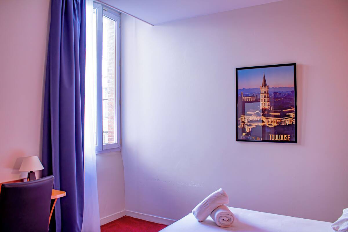 Een hotelkamer met een bed, een stoel, een tafel met een lamp, een raam met gordijnen en een stadsgezicht poster aan de muur.