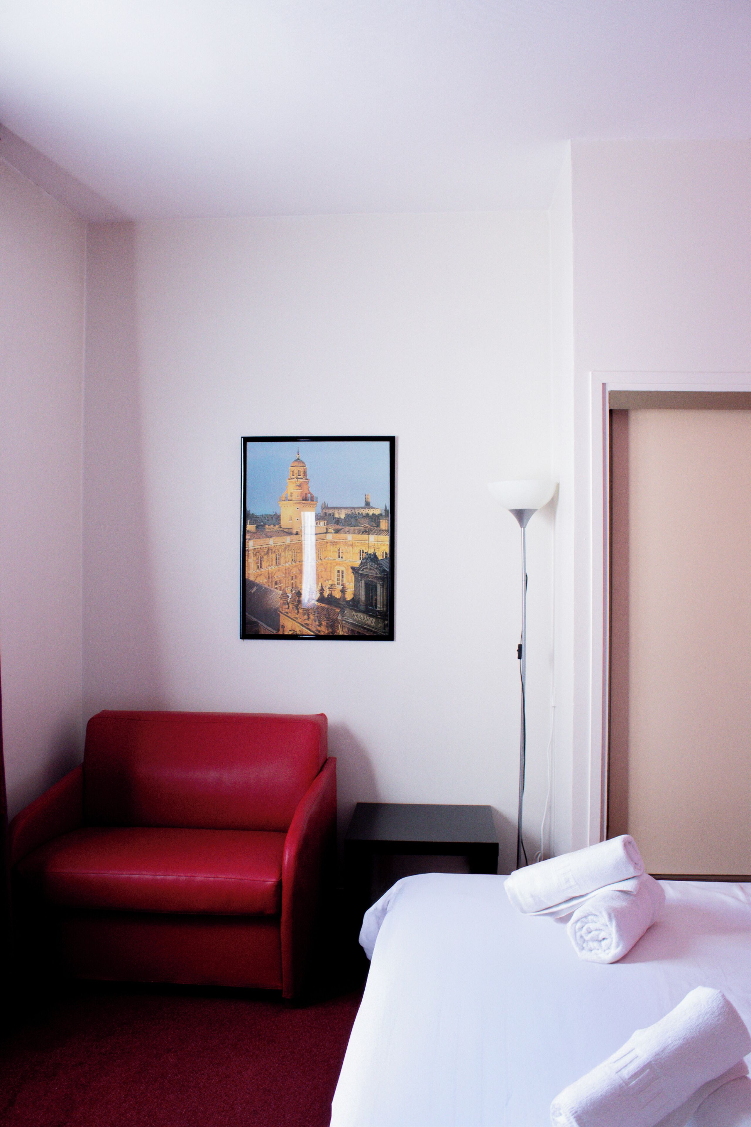 Een hotelkamer met een bed, een rode fauteuil, een nachtkastje, een lamp en een lijst met een afbeelding aan de muur.