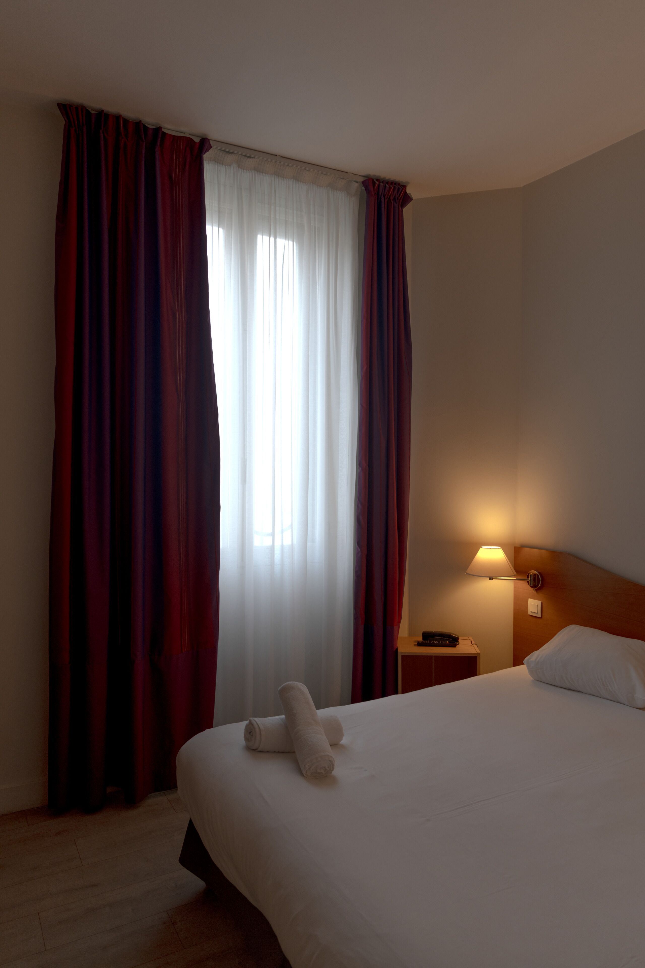 Una habitación de hotel con una cama, una mesita de noche, una lámpara y un ventanal con cortinas.